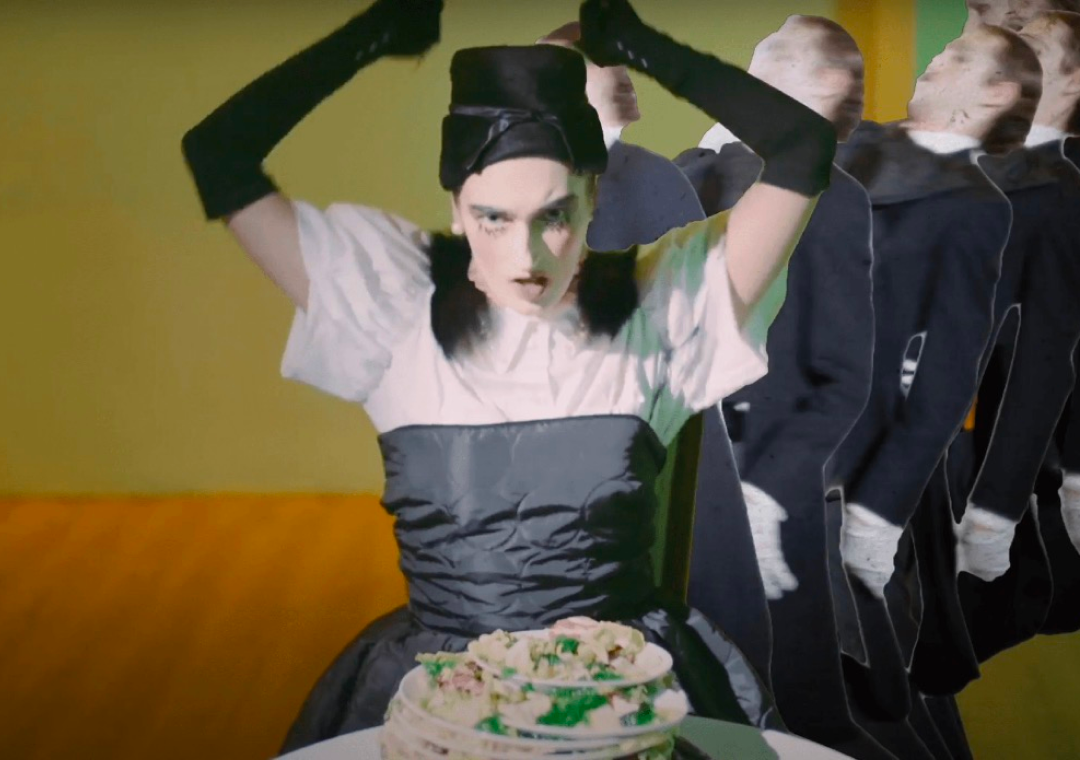 Mujer con expresión intensa y maquillaje oscuro, con ropa blanca y corset negro, lleva guantes largos negros, sentada junto a un pastel decorado con flores, en un ambiente con fondo de color verde y personas de espaldas a ella.