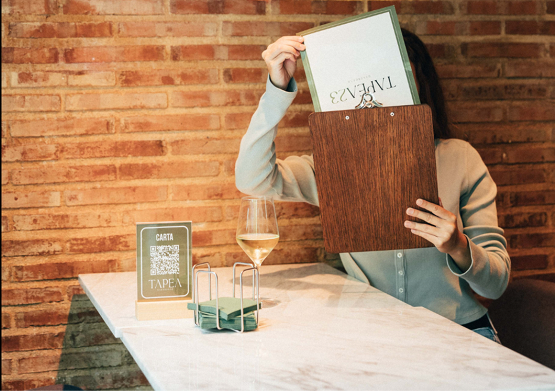 Persona sosteniendo el menú en un restaurante, con un vaso de vino y una mesa con señalización digital que dice 'Carta' con código QR.