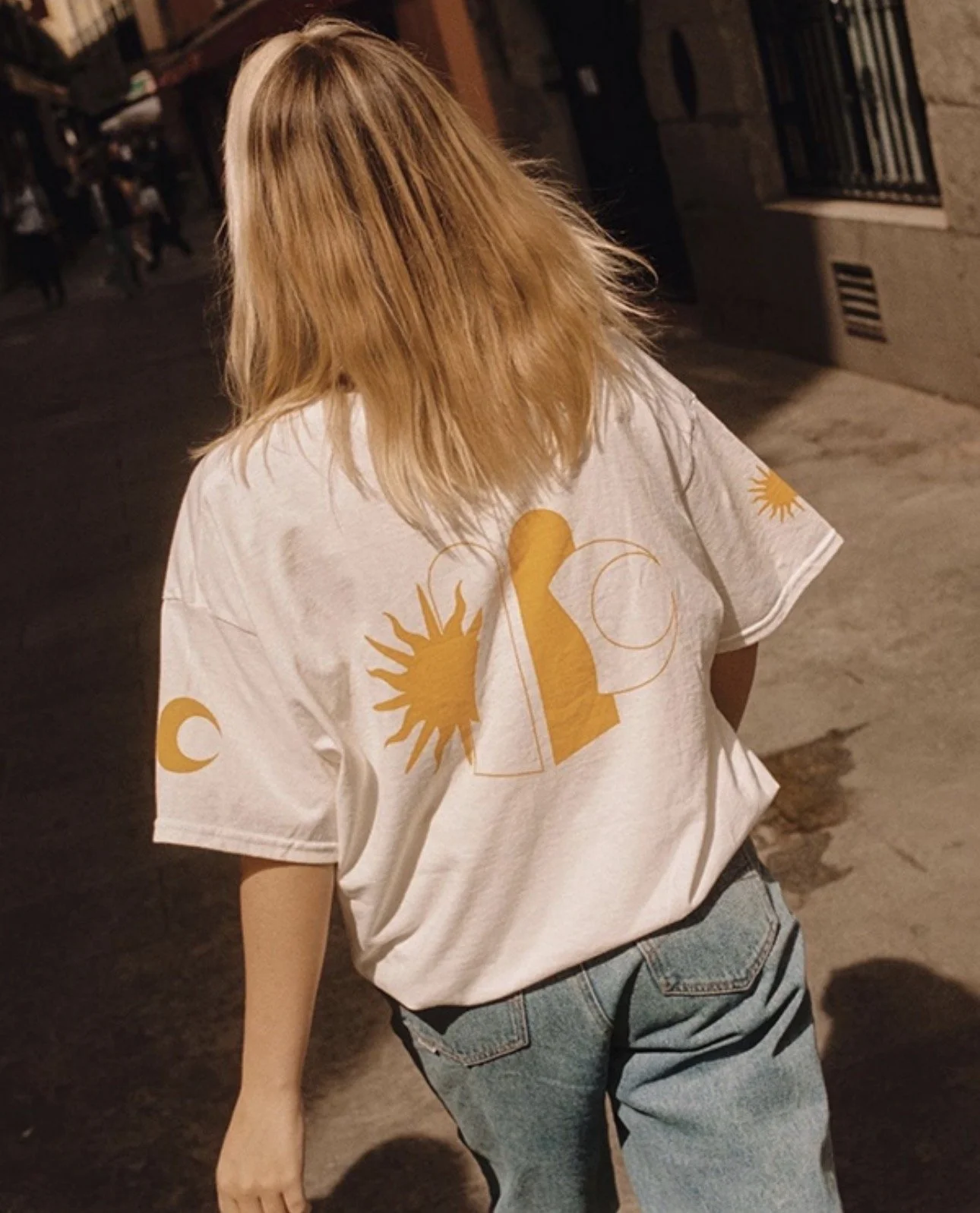 Una persona con cabello rubio y largo vista de espaldas, lleva una camiseta blanca con dibujos en color amarillo de un sol, una luna y una figura estilizada, y usa jeans azules, caminando por la calle de noche.