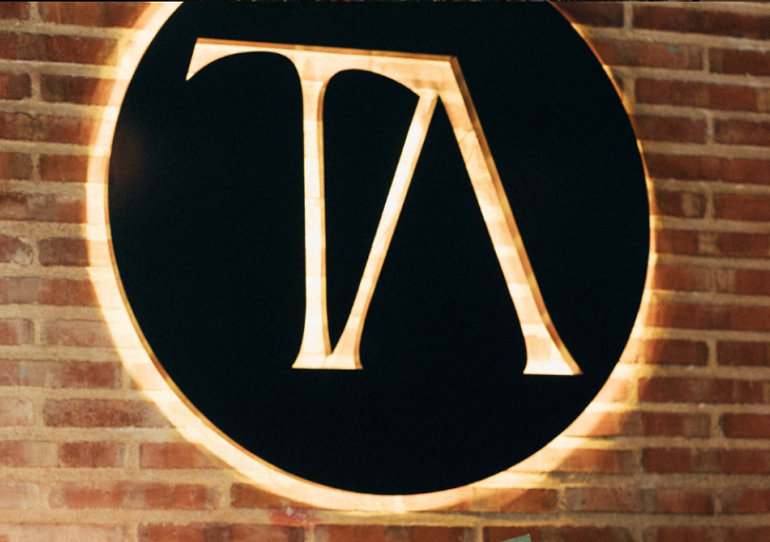 Logo de color negro con letras estilizadas 'V/A' en un círculo, en una pared de ladrillos.