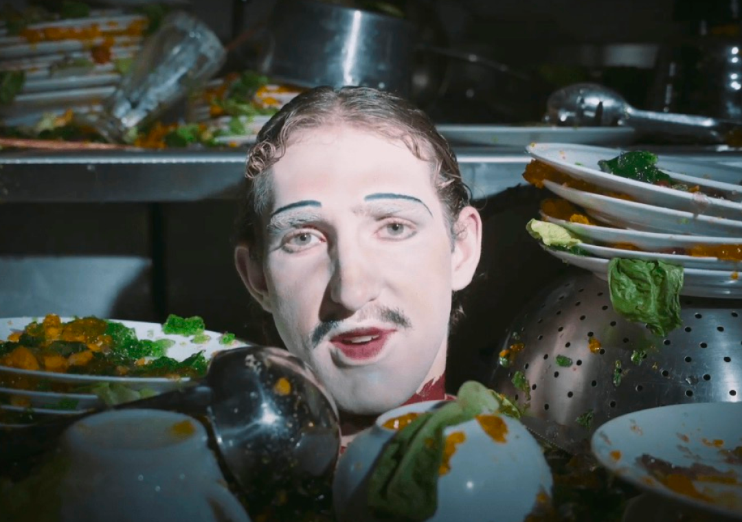Hombre con maquillaje de payaso entre platos y utensilios de cocina, rodeado de comida y platos sucios en una cocina.