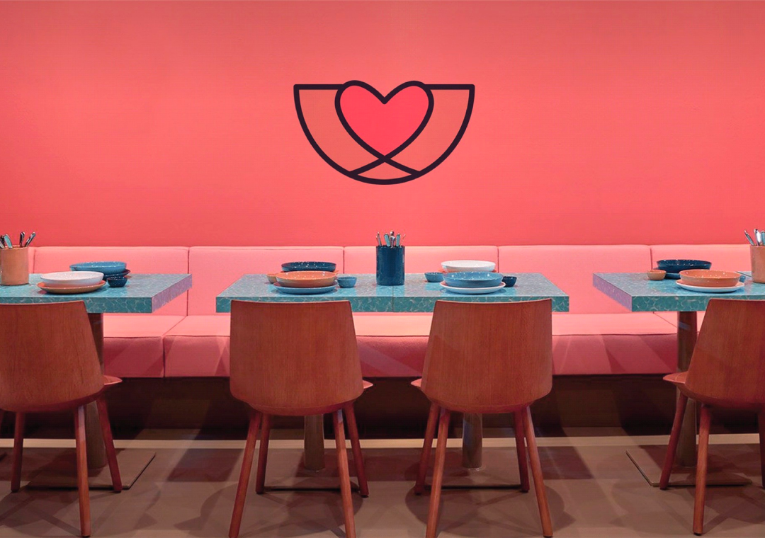Mesas de restaurante con platos y cubiertos en un lugar decorado en tonos rosa y madera, con un fondo de pared rosa y un símbolo de corazón y alas en la pared.