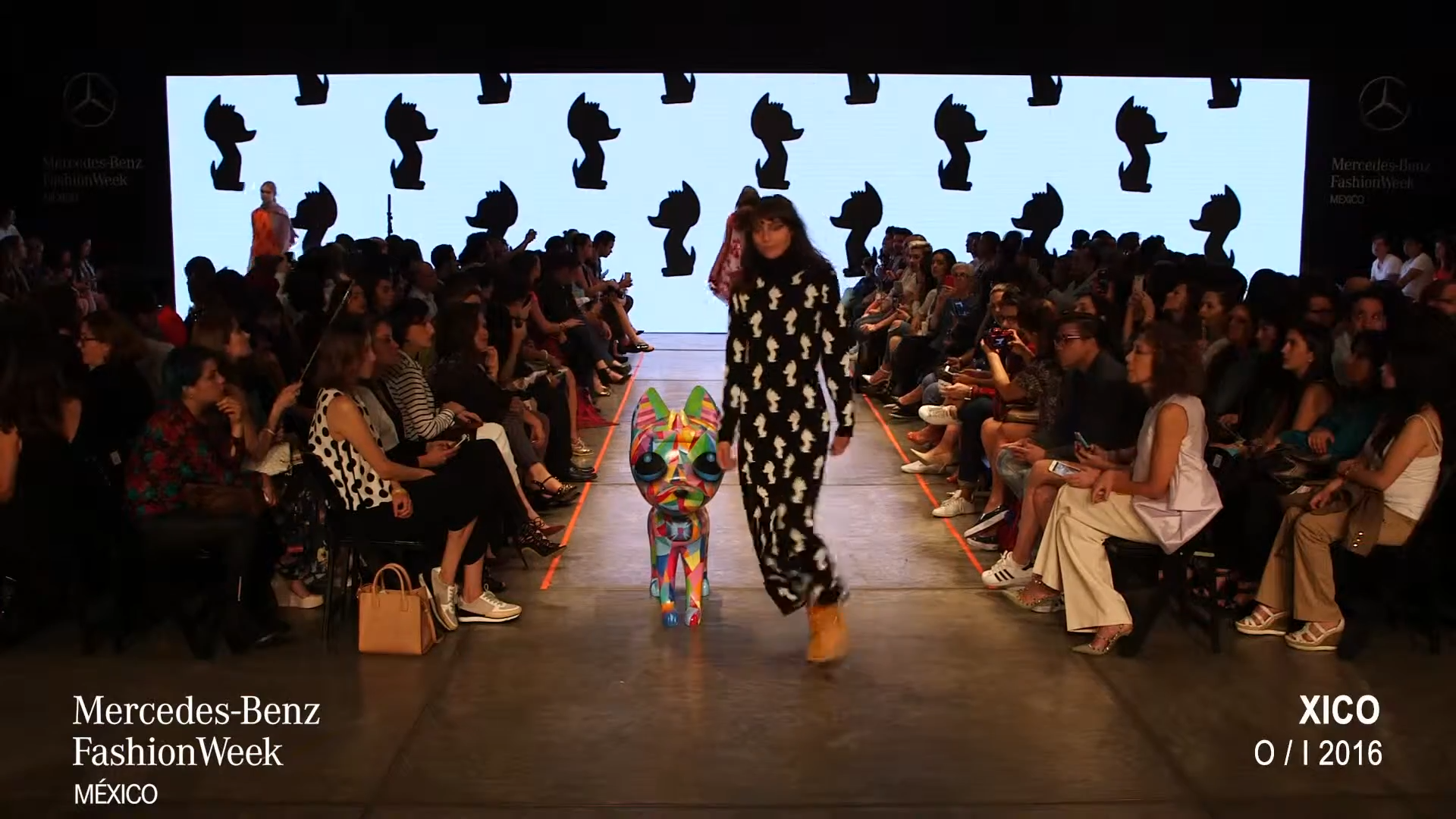 Pasarela de moda en el Mercedes-Benz Fashion Week en México con un público sentado a los lados, una modelo caminando y una pantalla grande con siluetas negras en fondo blanco.
