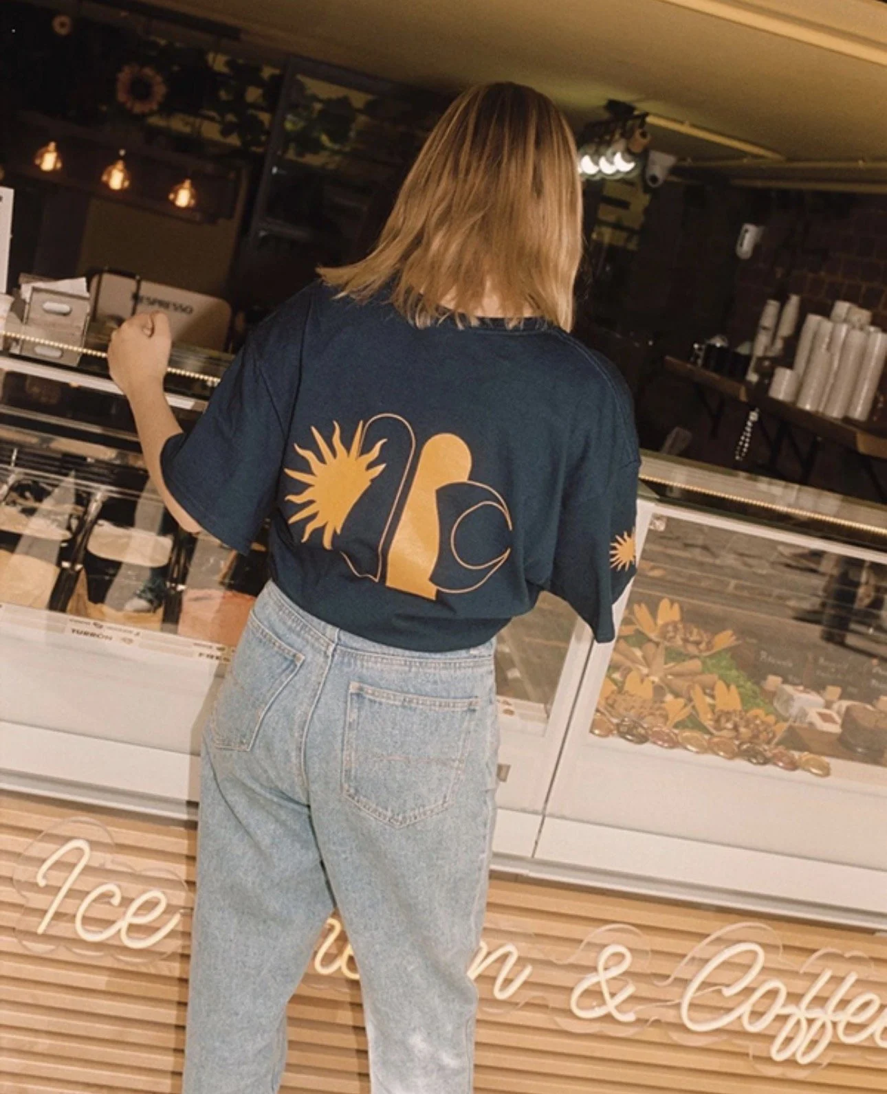 Mujer con blusa azul y jeans claros en heladería, con vitrina de helados y cartel de corazón y flores en fondo.