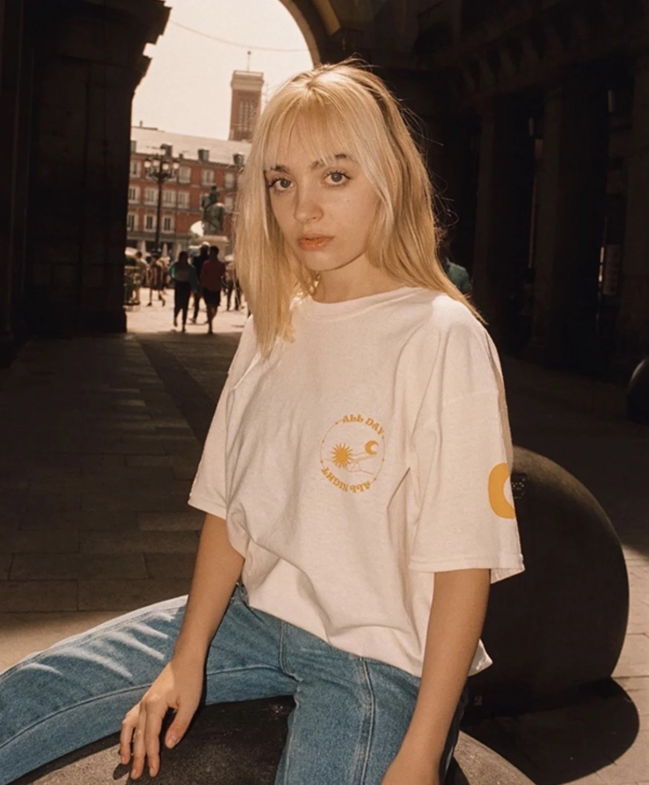 Joven mujer con cabello rubio y camisa blanca con diseño, sentada en una plaza bajo un arco, con edificios y gente en el fondo, luz cálida de la tarde.