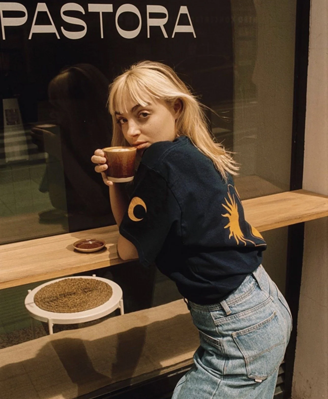 Una joven con cabello rubio y ropa oscura, sostiene una taza, frente a una ventana con textos en mayúsculas, en un ambiente de cafetería en la noche. Hay un plato con semillas y una pequeña bandeja con salsa sobre la mesa de madera.