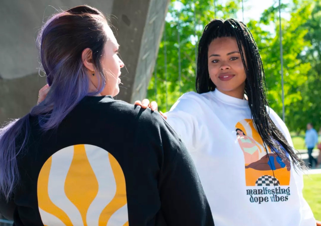 Dos mujeres conversando al aire libre, una lleva sudadera blanca con un diseño y texto, la otra sudadera negra con un logotipo grande en la espalda, en un parque con árboles verdes.