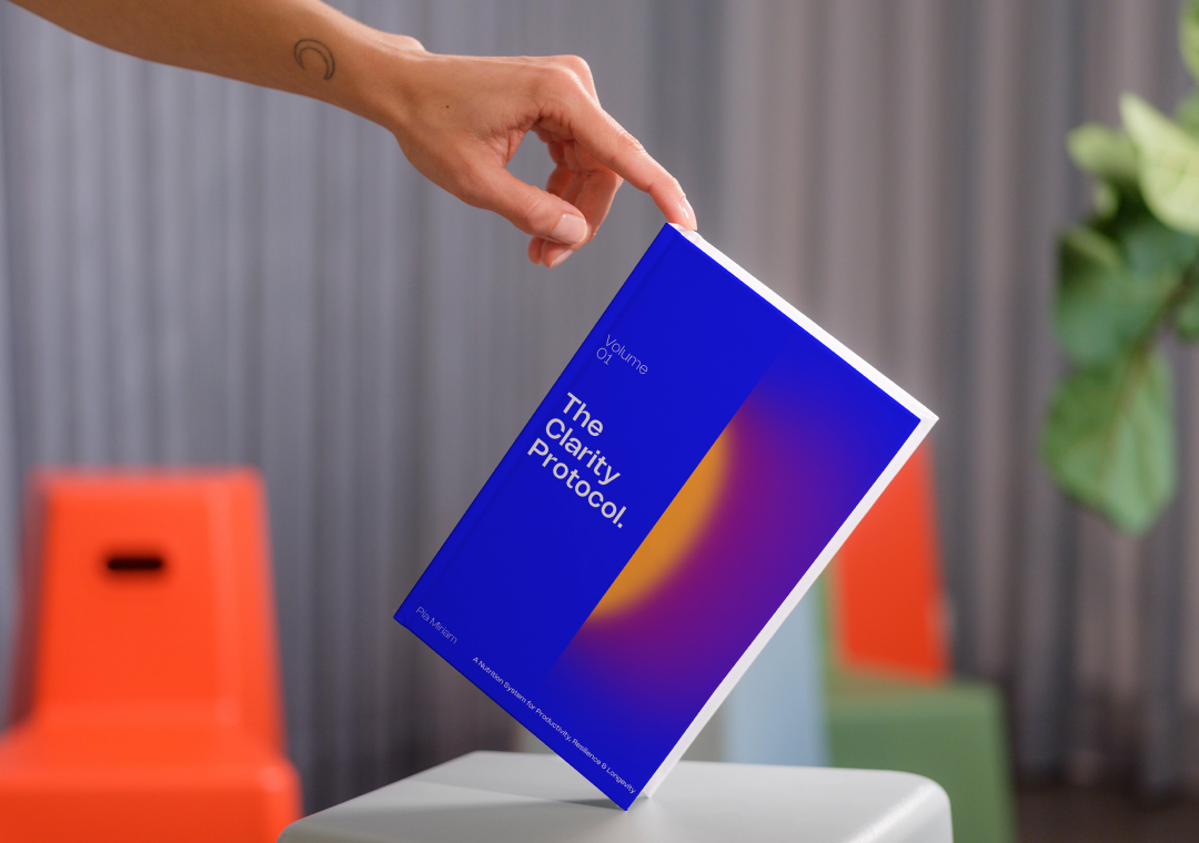 Una mano sosteniendo un libro titulado 'The Clarity Protocol' en un entorno interior con sillas de colores y una planta en el fondo.