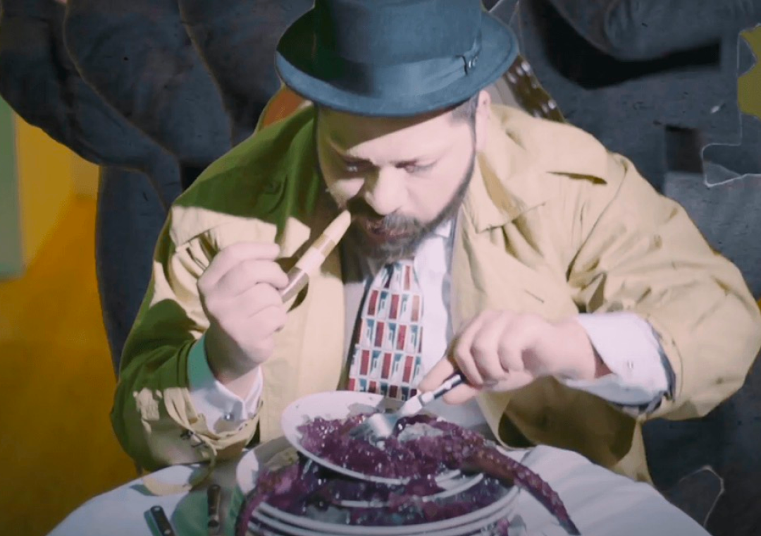 Hombre con sombrero negro, camisa con estampado de banderas y chaqueta beige, comiendo en un plato con lo que parece ser un molusco, en un entorno de comedor.