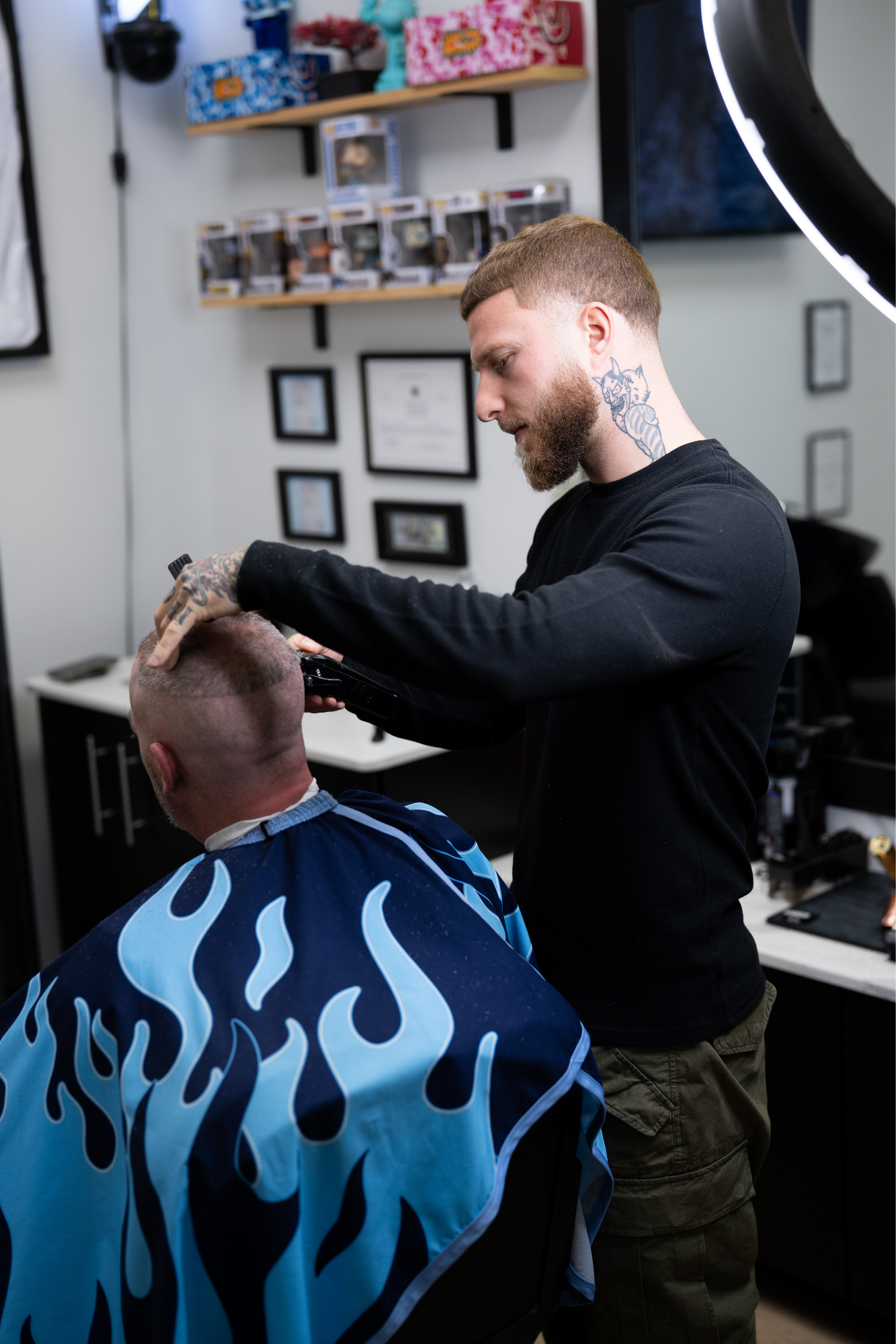 Barber cutting a customer's hair in a barber shop, with shelves of products and framed certificates on the wall in the background.