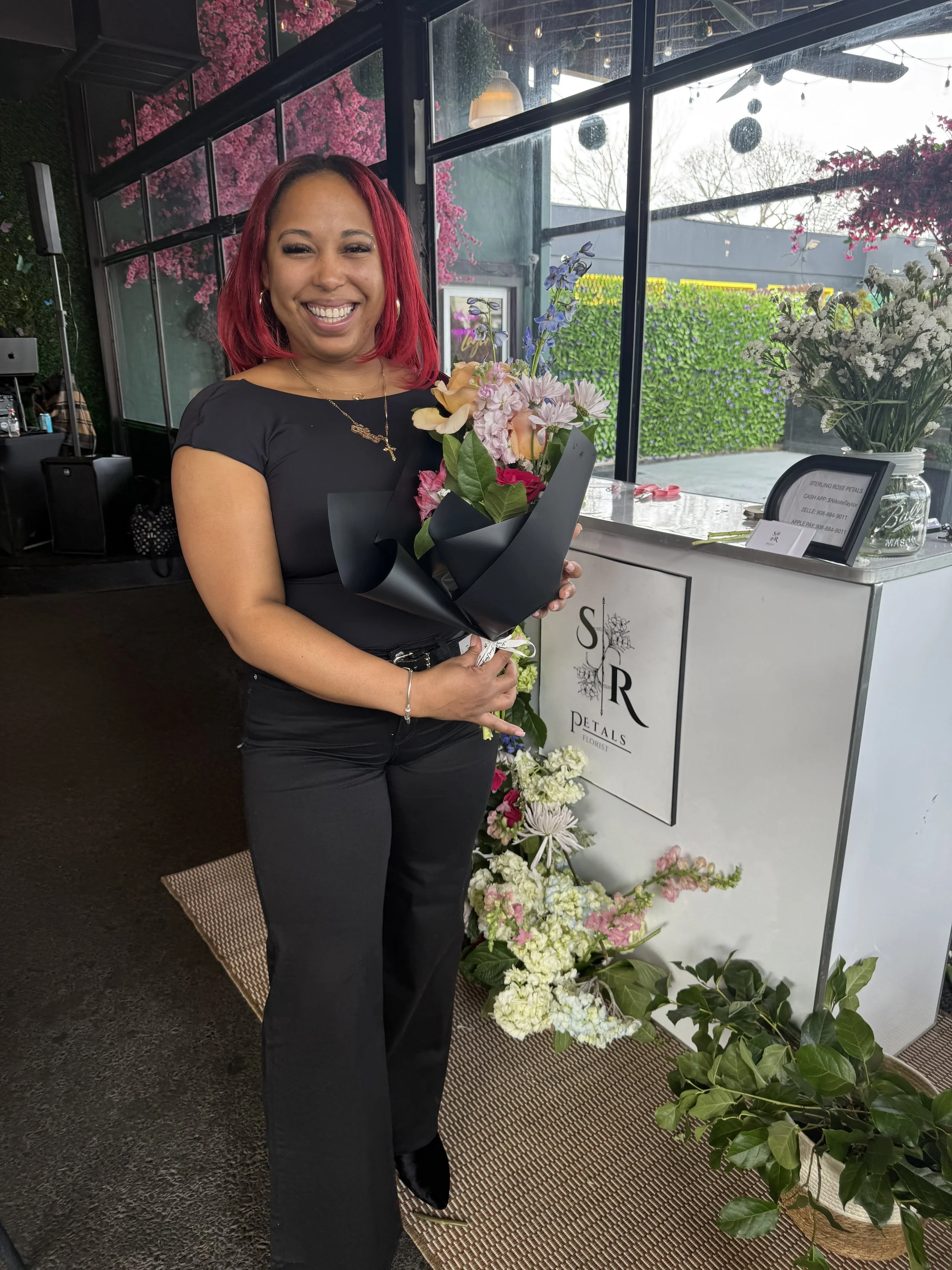 A woman with red hair standing inside a flower shop, holding a bouquet of pink, peach, purple, and red flowers. There are various floral arrangements and greenery around her, with a wall of pink blossoms behind her and a window showing outdoor trees and sky.