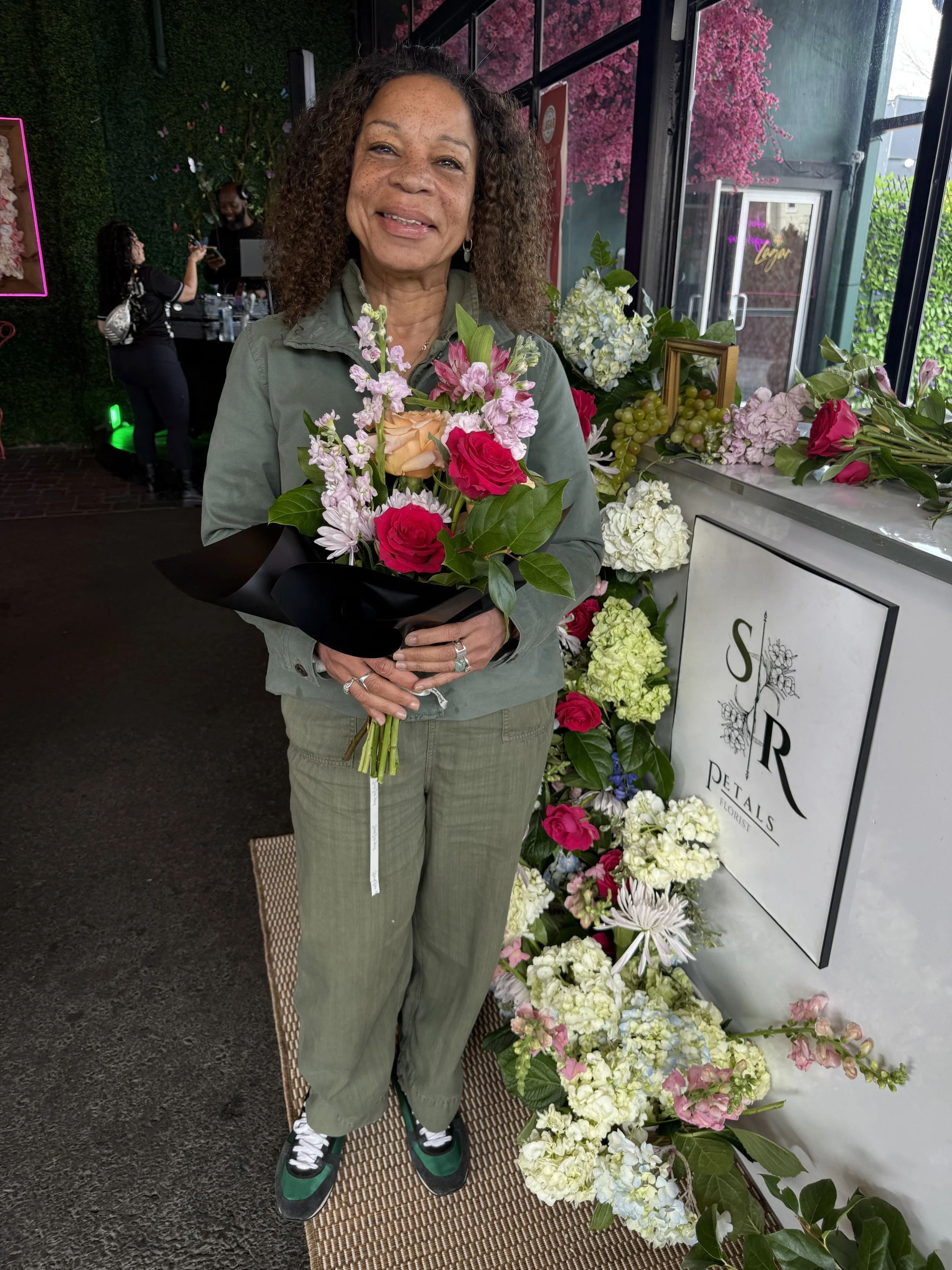 A woman with curly hair and freckles holding a bouquet of pink and peach roses and pink flowers, standing in front of a flower display at a florist shop.