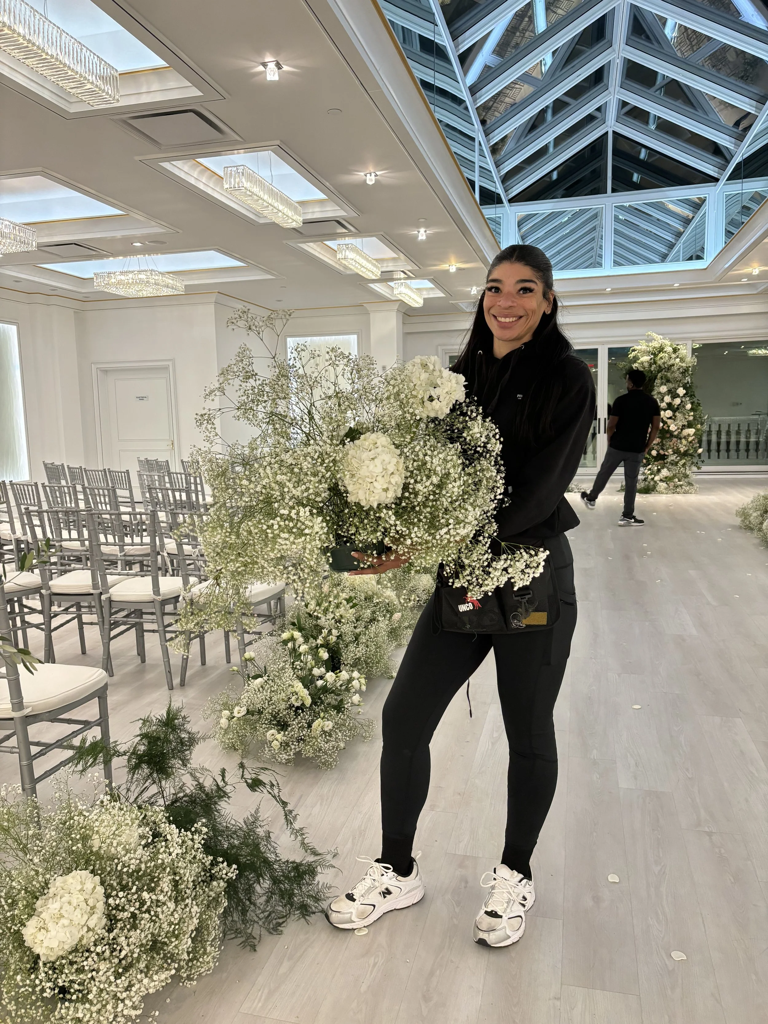A woman standing indoors holding a large bouquet of white flowers, smiling at the camera. She has black hair tied back and is dressed in a black hoodie, black pants, and white sneakers. The background features a decorated event space with rows of chairs, floral arrangements, and a glass ceiling.