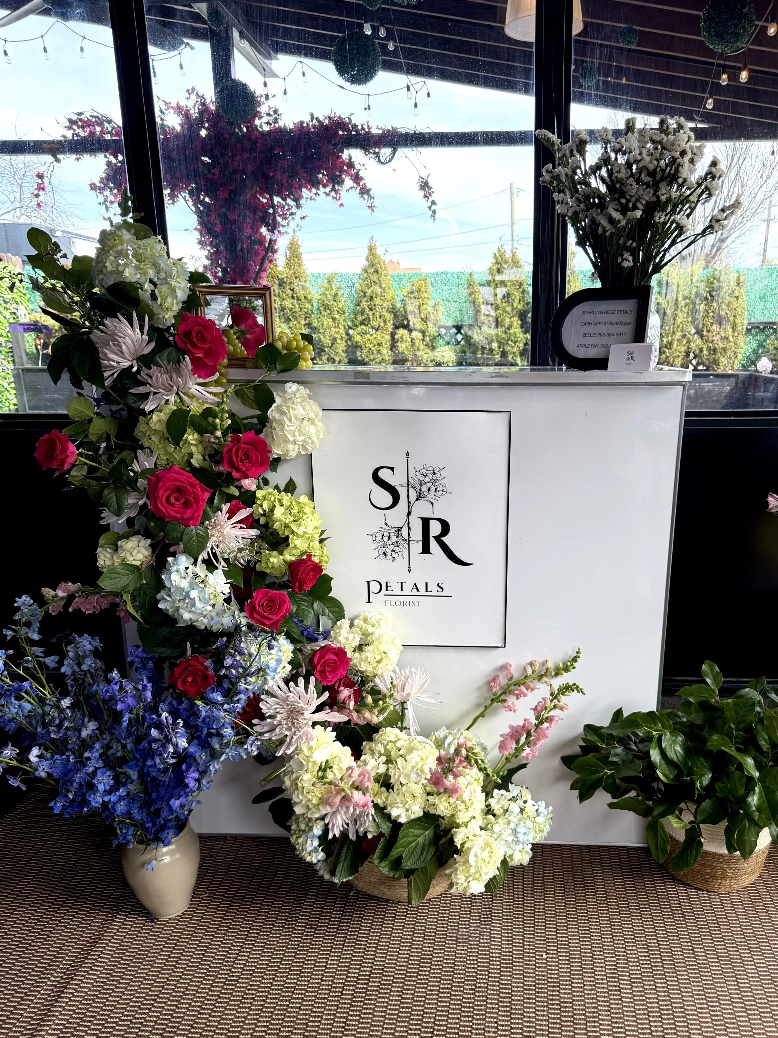 Arrangement of various colorful flowers including roses, hydrangeas, and other blooms displayed in vases and baskets, with a sign for a florist shop behind them, in front of a large window with outdoor trees and sky visible.