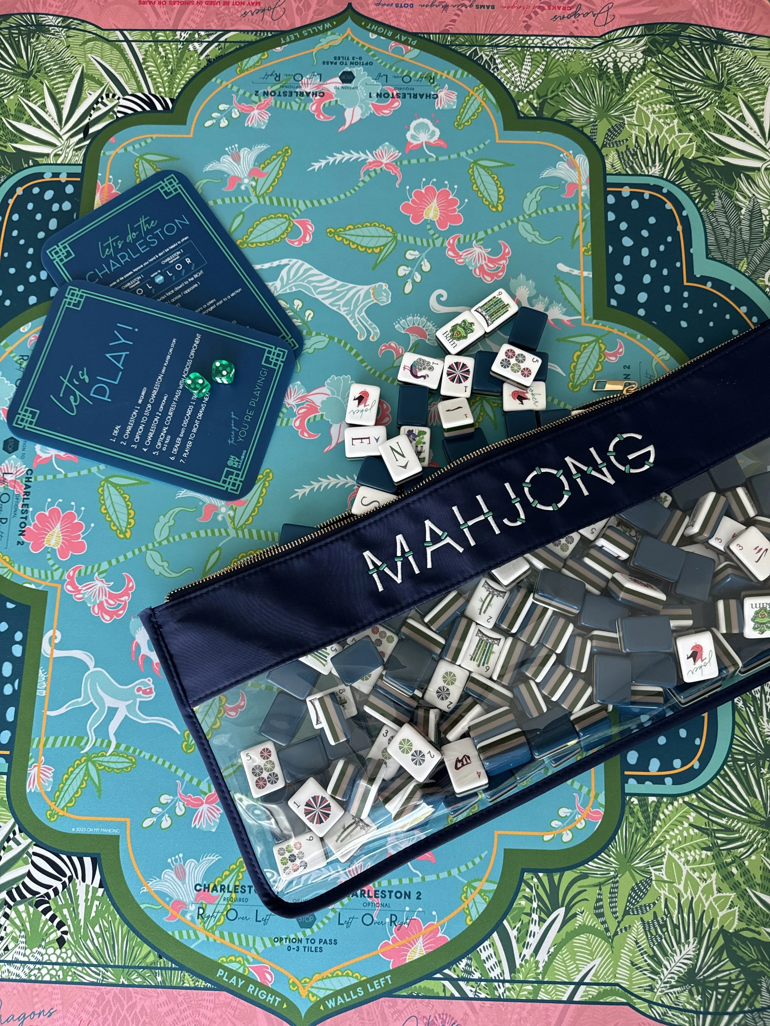 A decorated board game with various game pieces, cards, and a navy blue case on a colorful, patterned surface featuring lush green and pink tropical foliage and flowers.