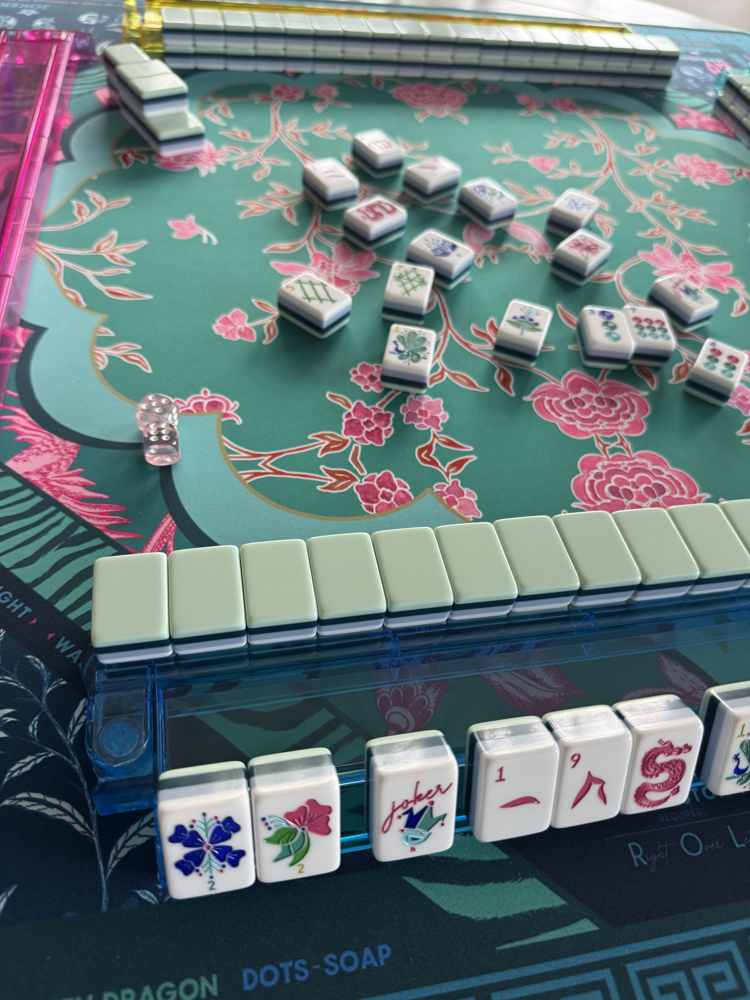 Close-up of a Mahjong game in progress on a floral tablecloth, with various Mahjong tiles arranged, a set of white tiles forming a wall, and some tiles with floral and decorative patterns visible.