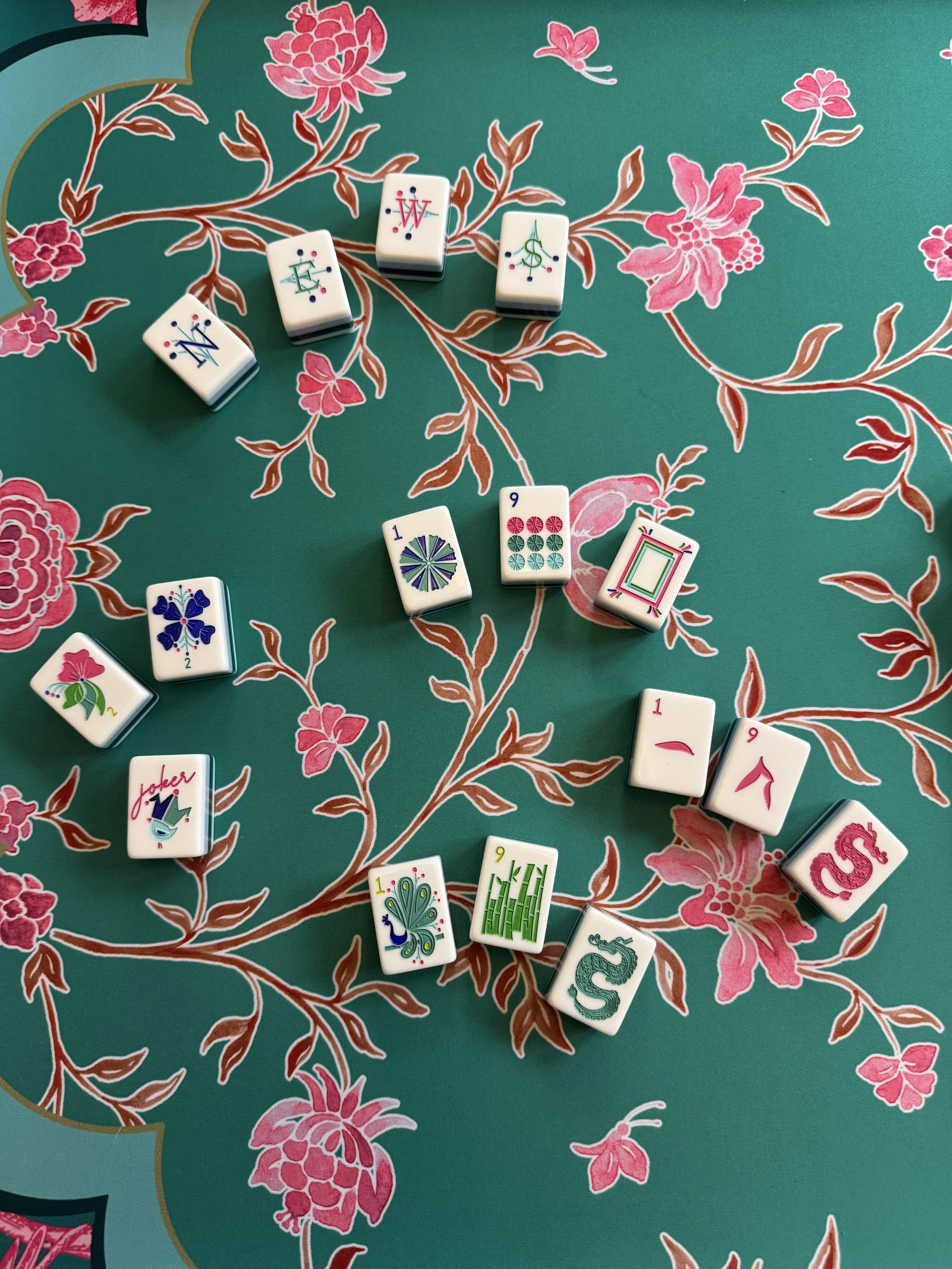Colorful Mahjong tiles arranged on a floral patterned tablecloth with pink and green flower designs.