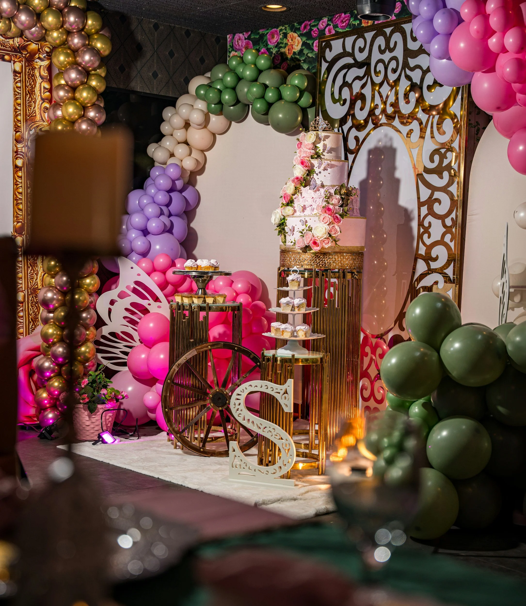 Decorative party backdrop with balloons in pink, purple, green, white, and gold, alongside a pink tiered cake adorned with flowers and a crown topper, and a cotton candy machine in the foreground.