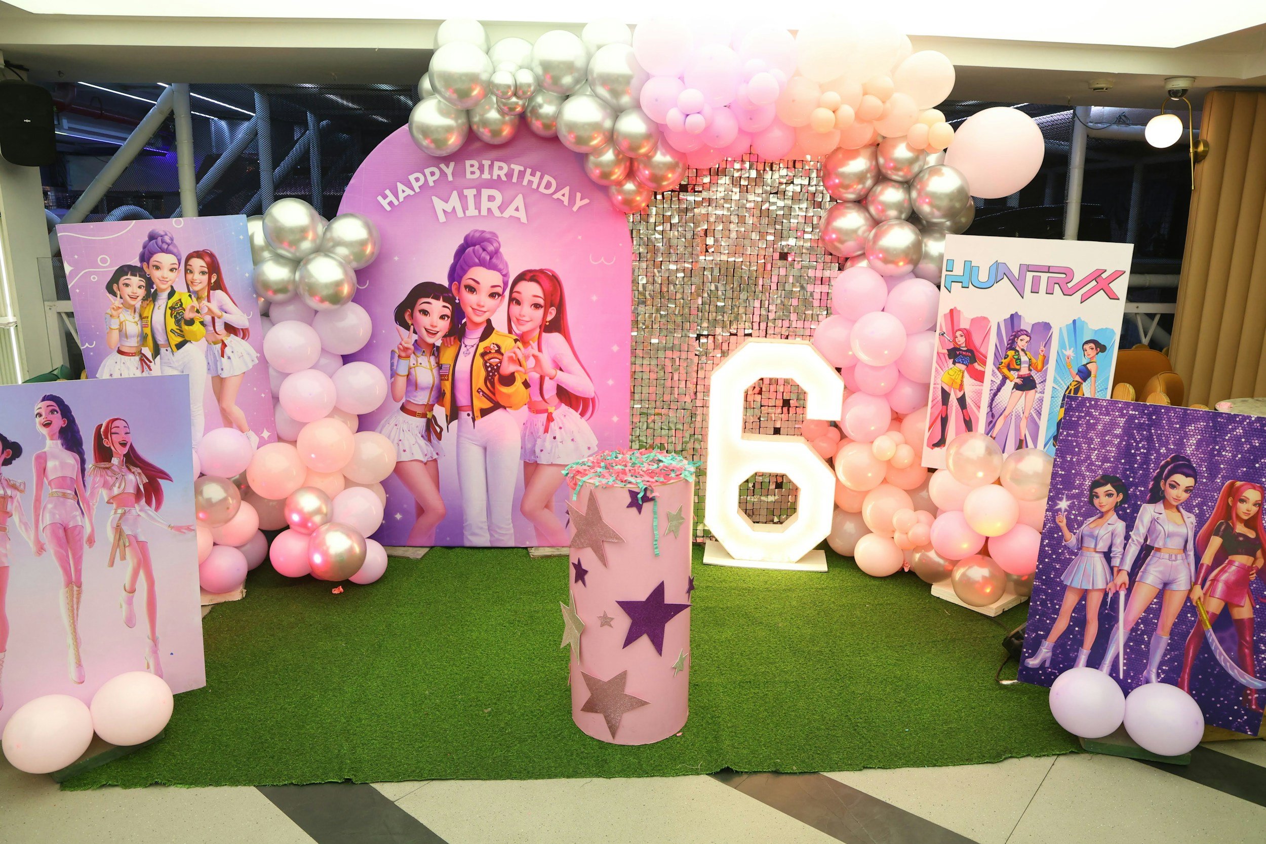 A decorated birthday celebration area with pink, white, and silver balloons, cartoon posters of girls, and a pink backdrop with the message "Happy Birthday Mira." There is a pink cake with teal and purple decorations on a table in front of the backdrop.