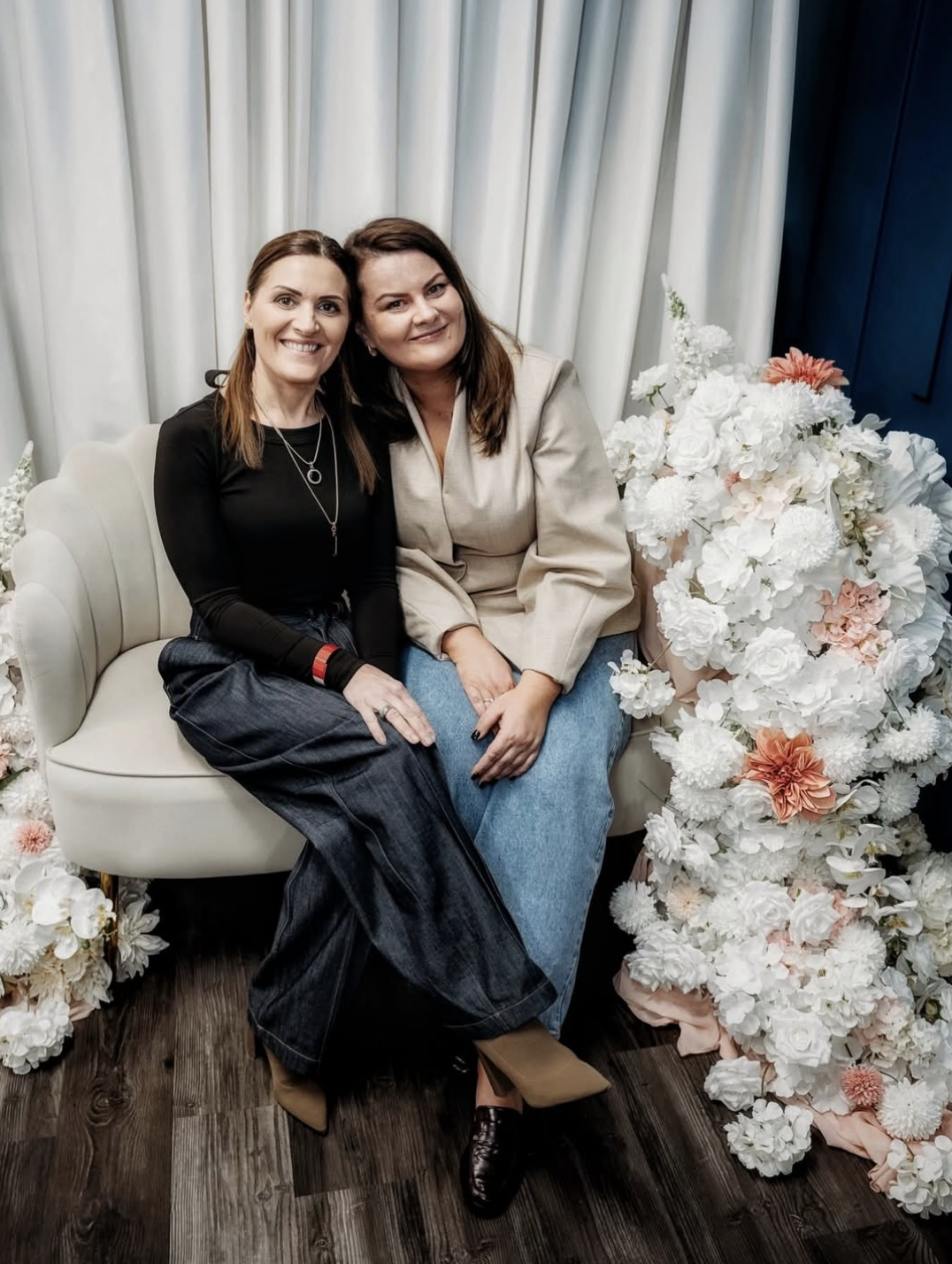 Two women sitting on a white sofa, surrounded by white and pink flowers, in front of white curtains, smiling at the camera.