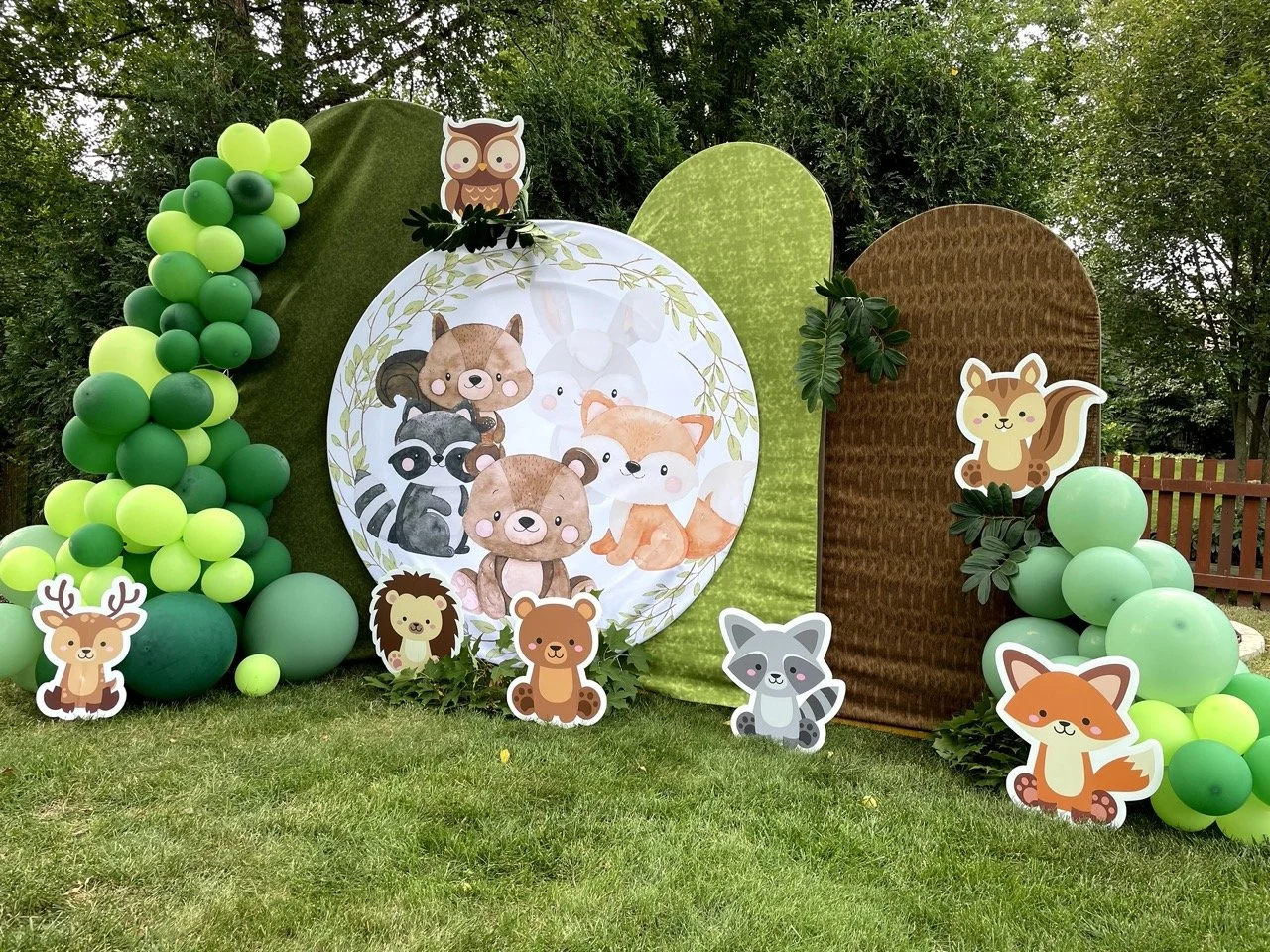 Decorative outdoor scene with cartoon animal cutouts including a deer, fox, raccoon, squirrel, bear, owl, lion, and others, arranged with green balloons, foliage, and fabric panels on a grassy lawn surrounded by trees.