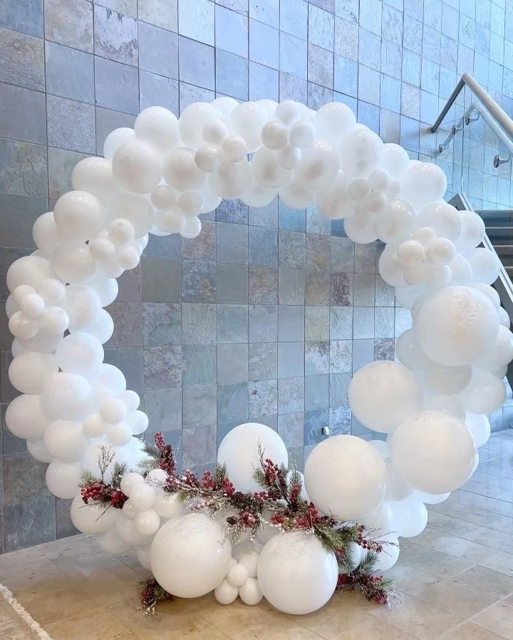 A large circular balloon arch made of white balloons of various sizes, decorated with sprigs of greenery and red berries at the base, set against a tiled wall near a staircase.