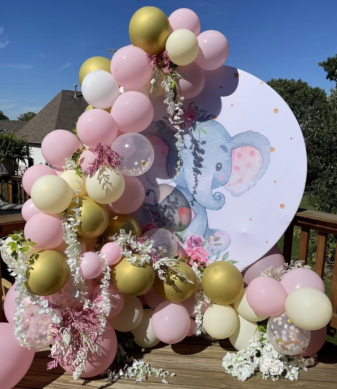 A decorated outdoor display featuring a large, round backdrop with a watercolor illustration of a blue elephant with pink ears, surrounded by pink, white, and yellow balloons and white floral arrangements for a baby shower or birthday celebration.