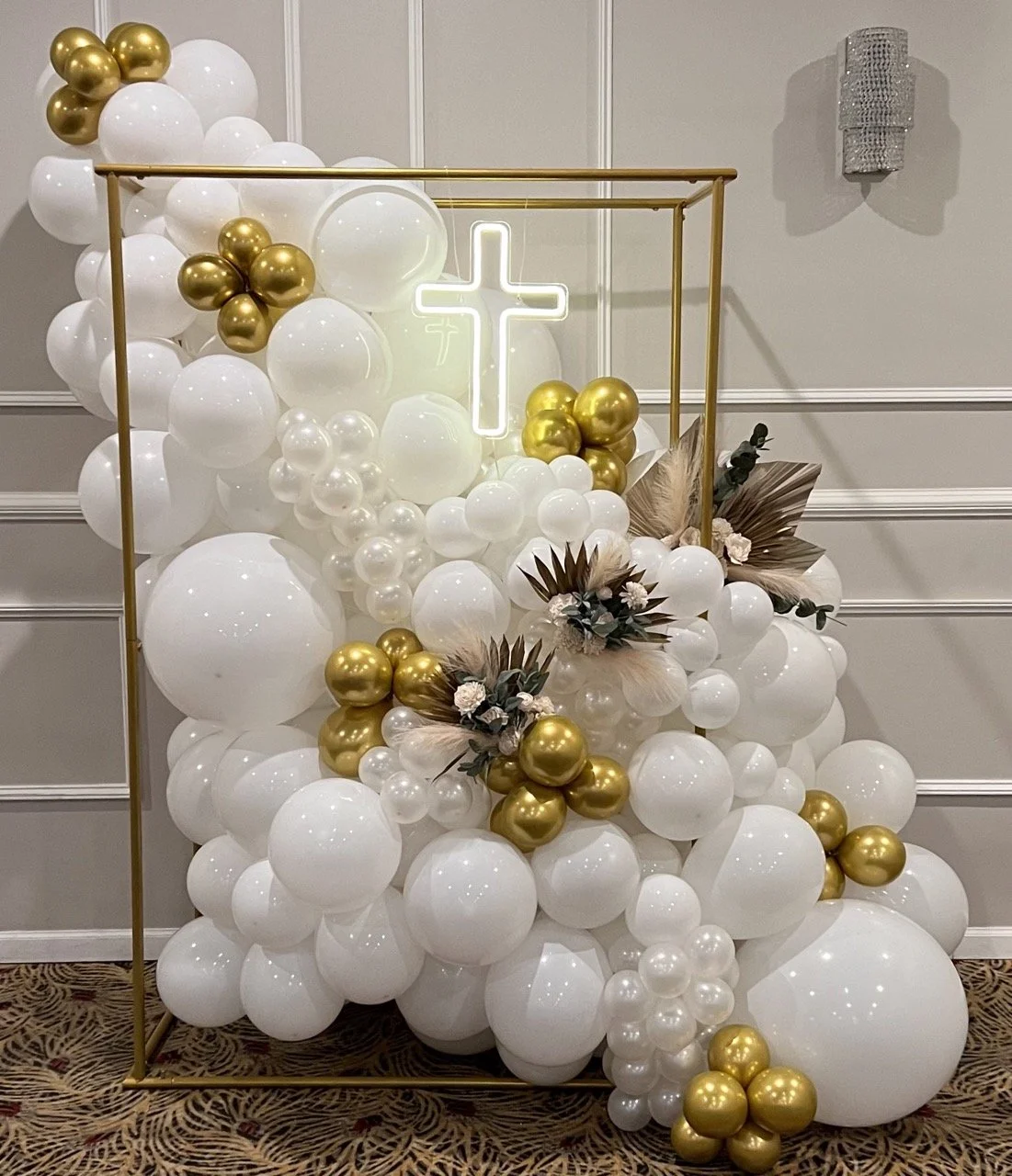 A decorative balloon display with white, gold, and pearl-colored balloons arranged inside a gold geometric frame, featuring a glowing cross neon sign and tropical leaves with blush-colored flowers.