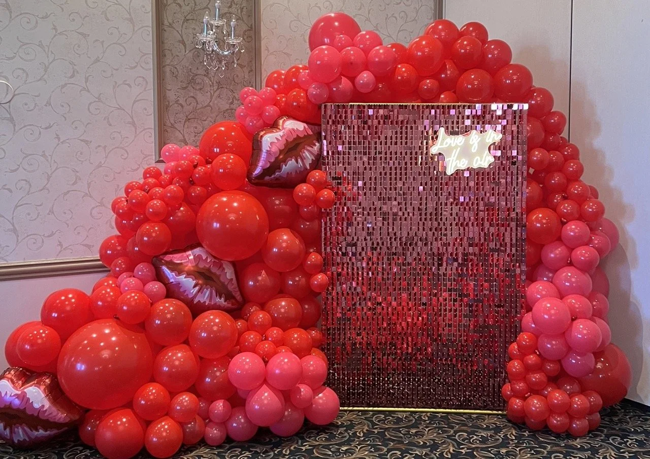 A decorative backdrop made of numerous red and pink balloons, with some lip-shaped balloons, set against an ornate wall with a chandelier. There is a shimmering mosaic panel with a neon sign that reads "Love & in the air."