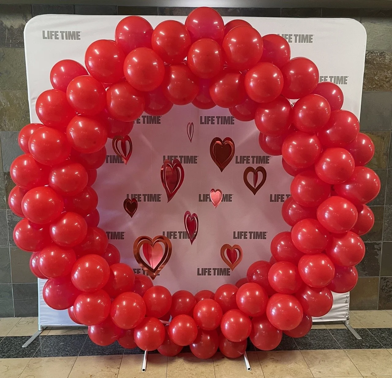 Red balloon arch in front of a backdrop with Heart shapes and the words "LIFE TIME" repeated multiple times.