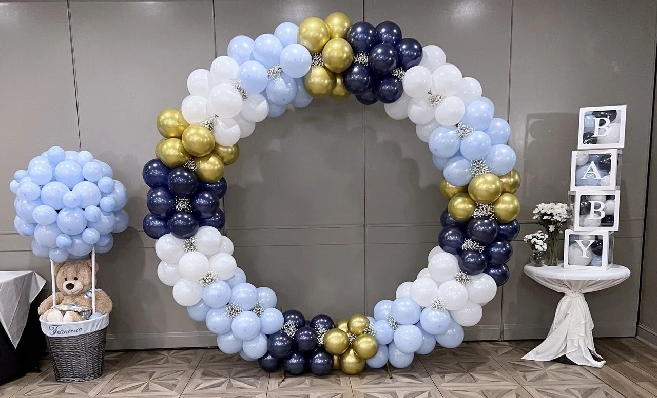 Decorative balloon arch in shades of blue, white, black, and gold with a balloon cluster and a display with framed letters spelling 'BABY' on a table with flowers and a teddy bear.