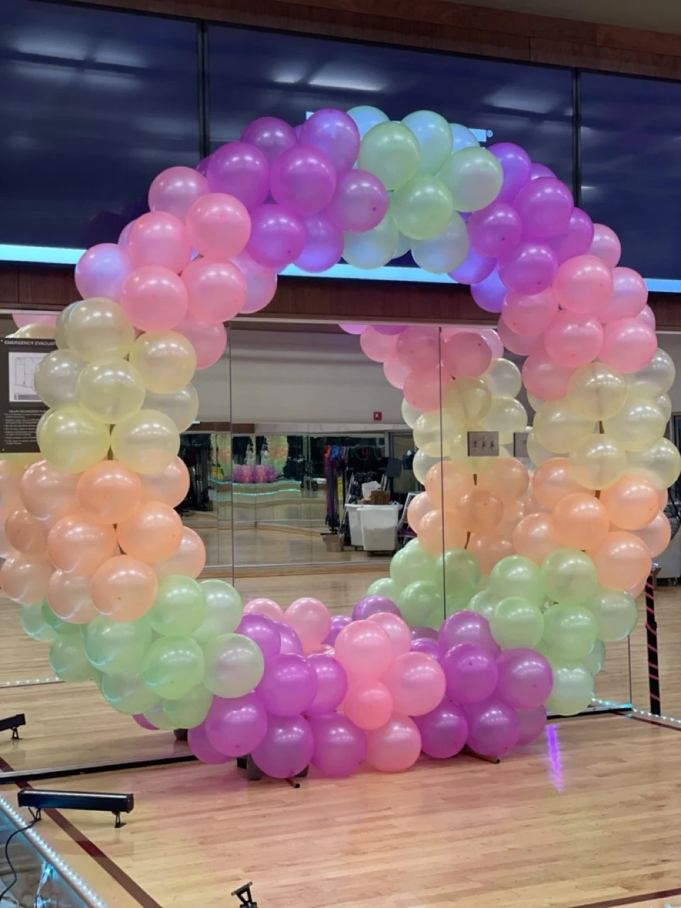 A balloon arch made of pastel pink, yellow, purple, and green balloons, decorated in a rainbow shape, situated in an indoor setting with a wooden floor and with mirrored panels in the background.