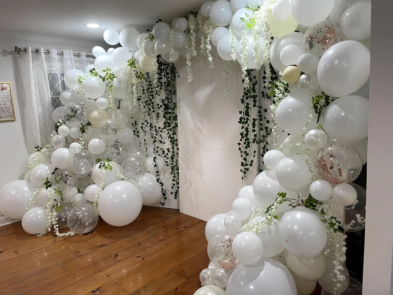 White balloon decoration with greenery and hanging white flowers on a wall, set up for an event or celebration in a room with wooden floor.