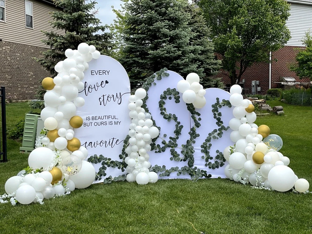 Outdoor wedding or celebration decoration with white and gold balloons, greenery, and a large white sign showing the initials 'J' and 'W' surrounded by leaves.