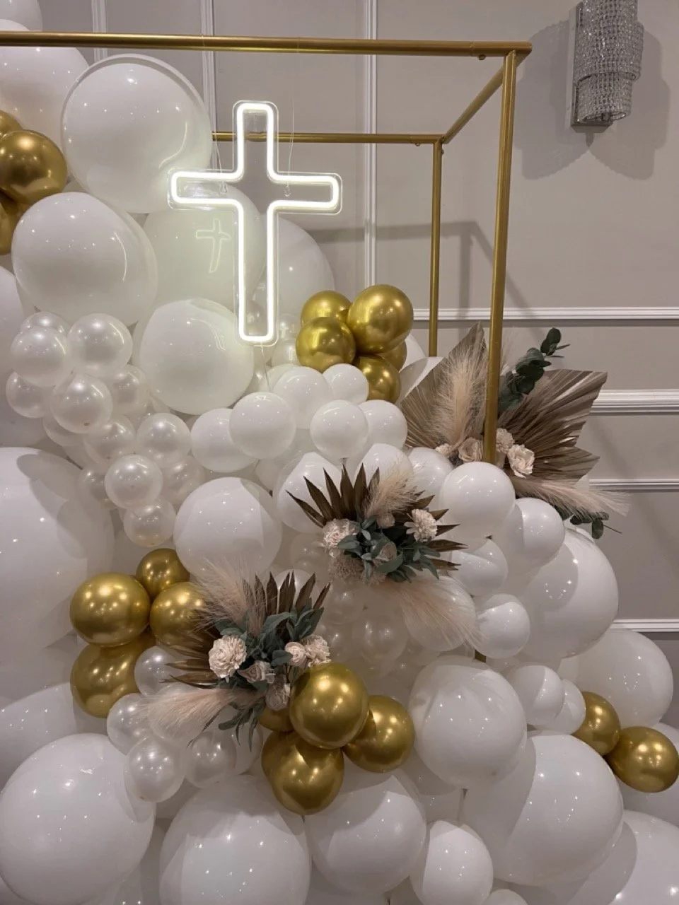 Decorative balloon arrangement with white, gold, and beige balloons, floral accents, a neon cross, and a gold frame.