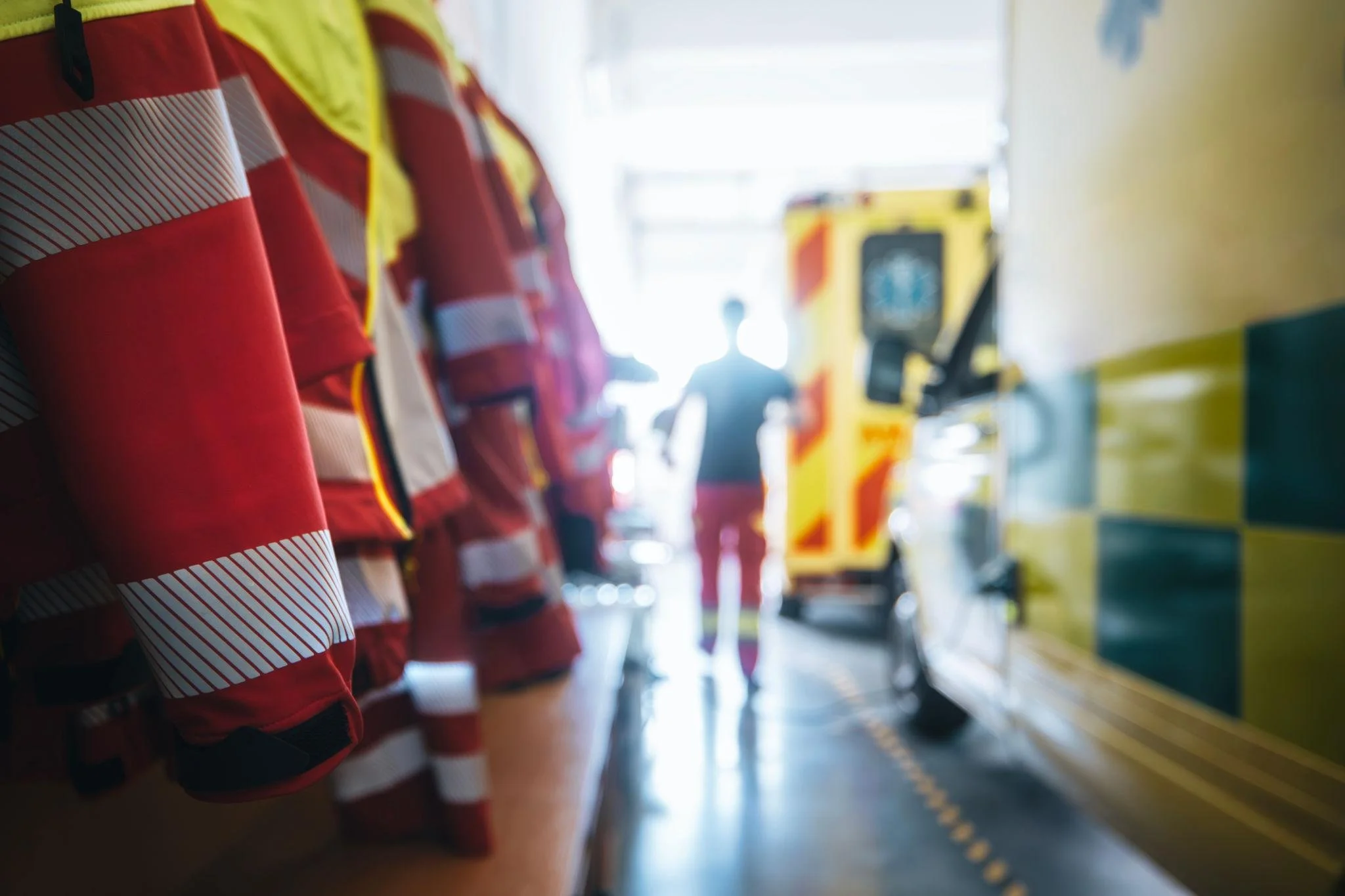 Lineup of firefighters in red and black gear at a fire station, with a yellow ambulance in the background.