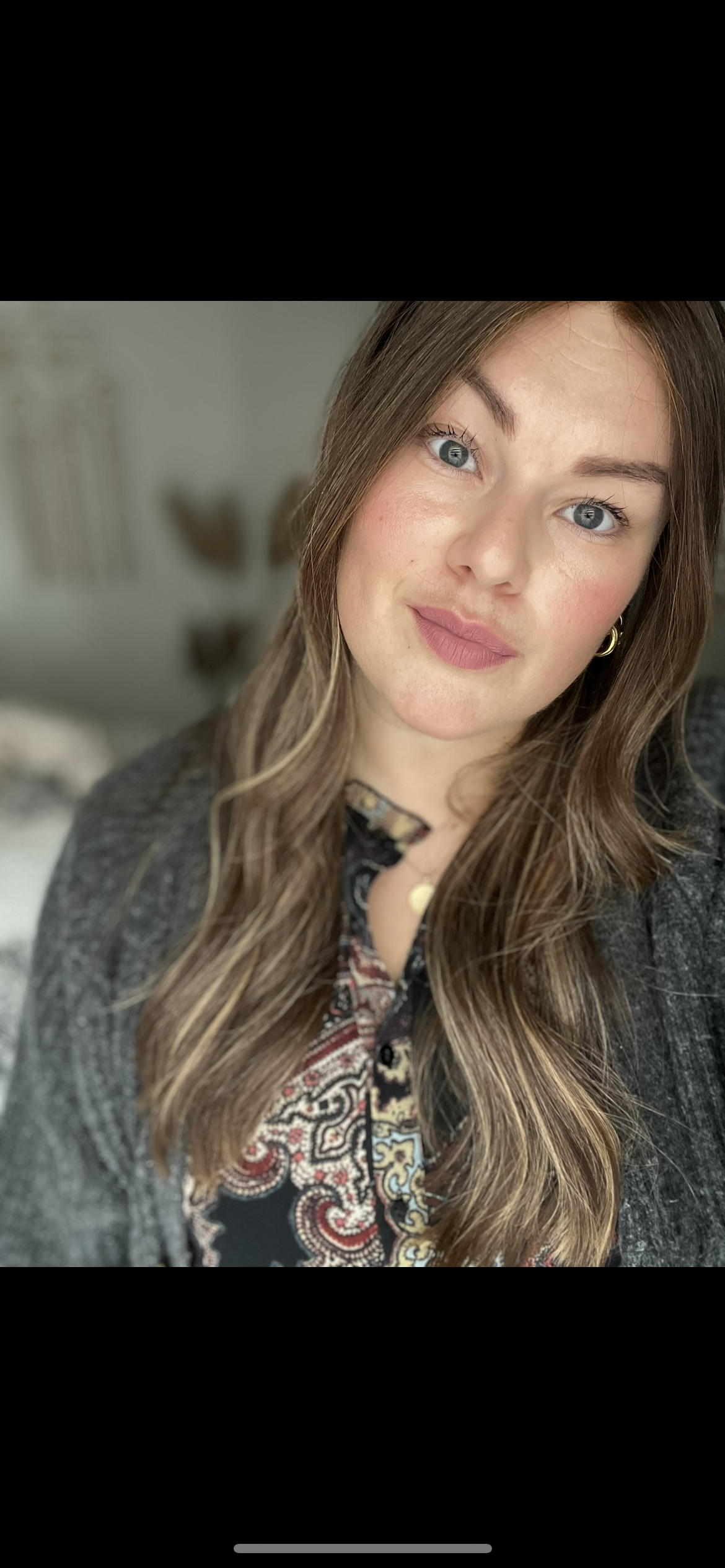 Une femme avec de longs cheveux bruns, des yeux bleus et un maquillage léger, portant un haut à motifs colorés et un cardigan gris, prenant un selfie dans une pièce intérieure.