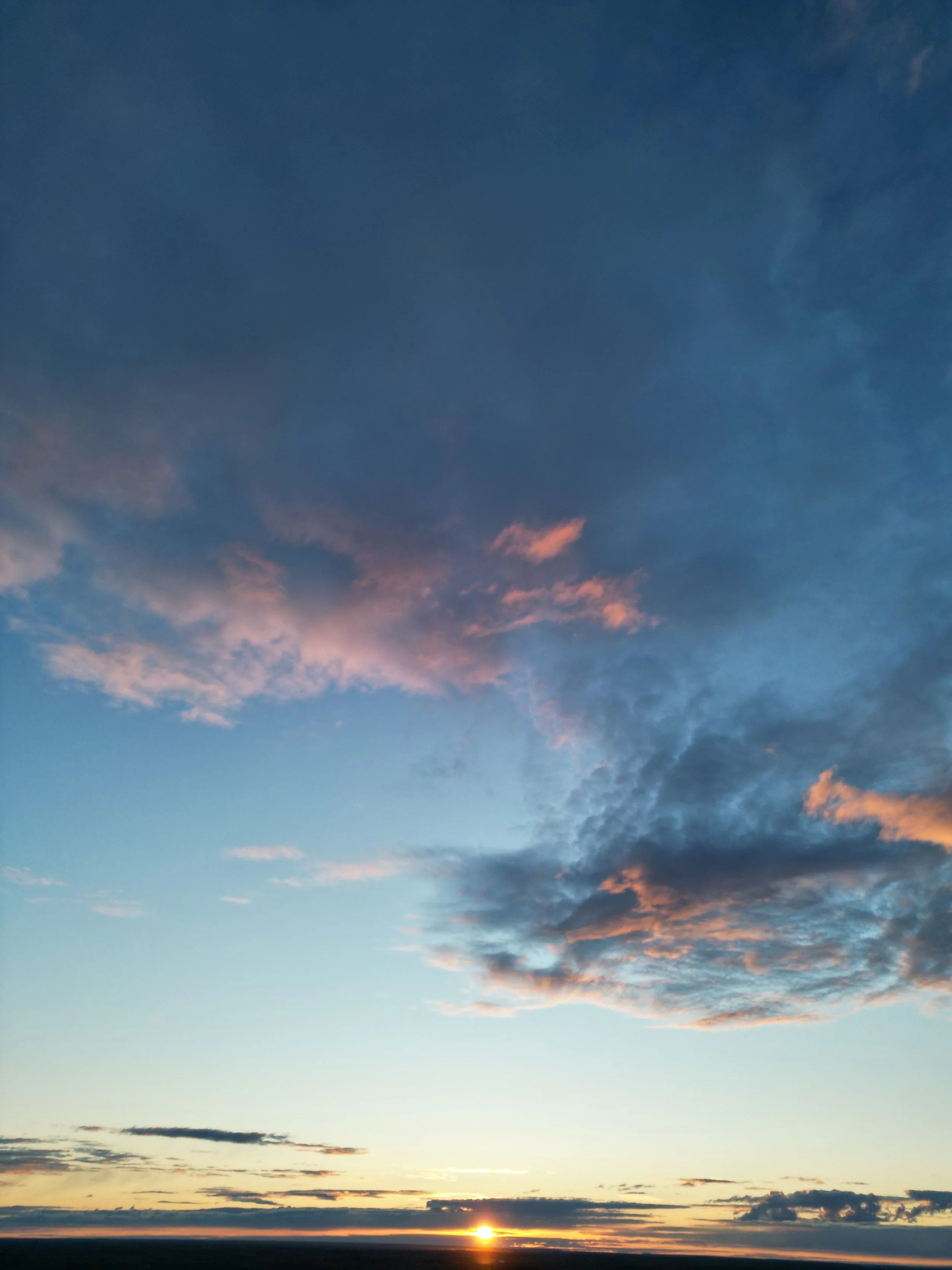 A sunset over a flat horizon with a dark sky and scattered clouds, some tinged with pink and orange hues.