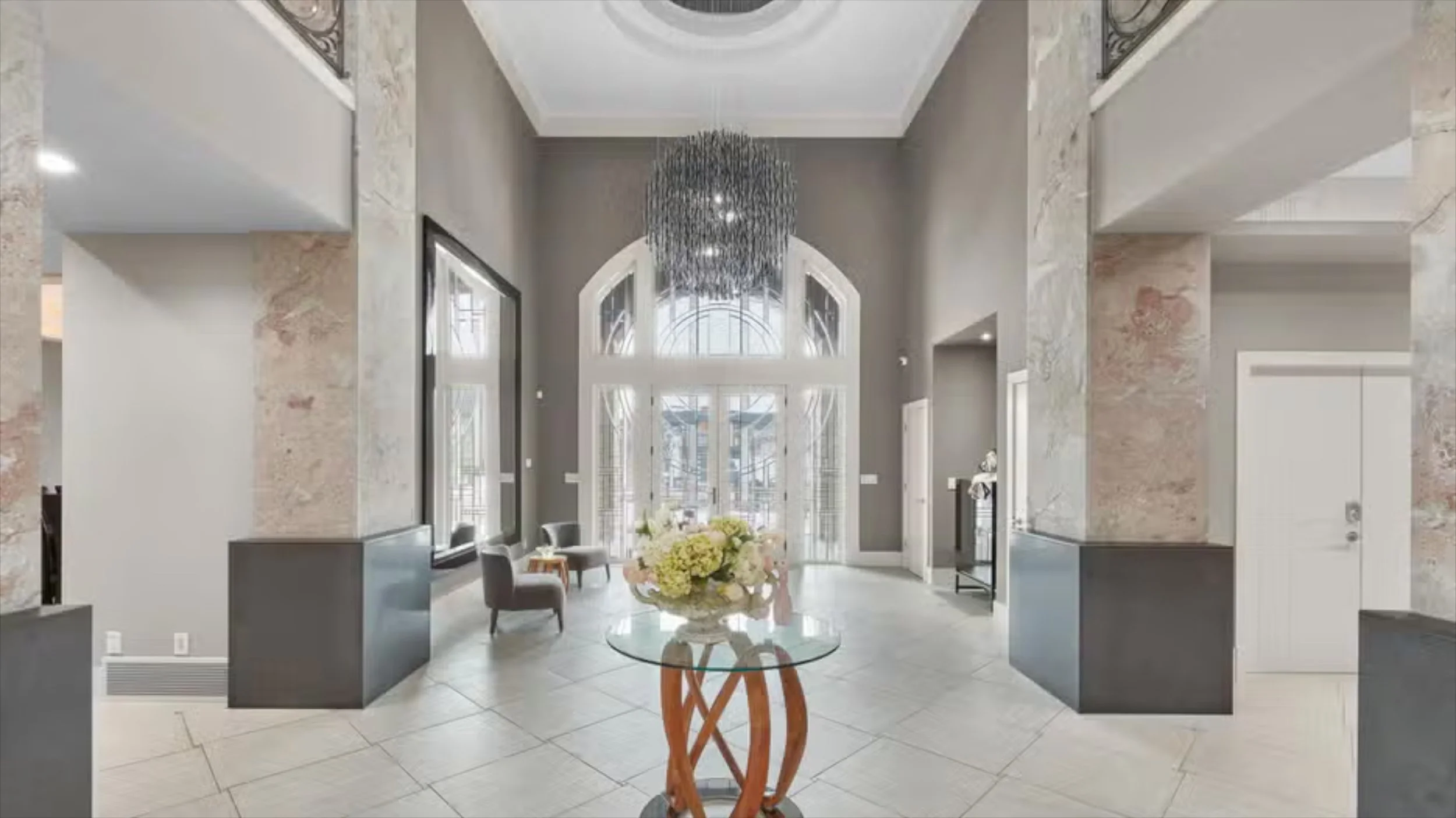 Elegant interior space with tall ceilings, large arched windows, and a modern chandelier, featuring a round glass table with a floral centerpiece and seating area.