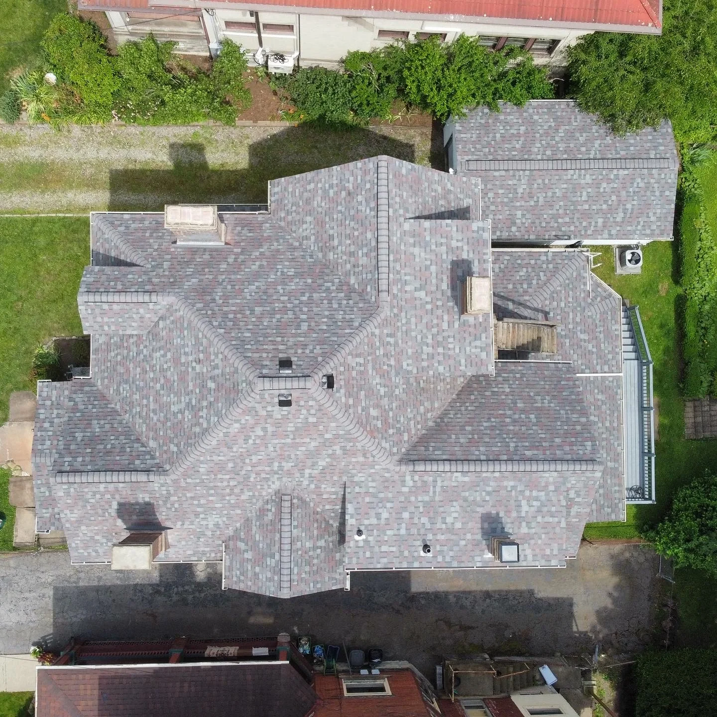 Aerial view of a house with a multicolored shingle roof, surrounded by greenery and neighboring buildings.