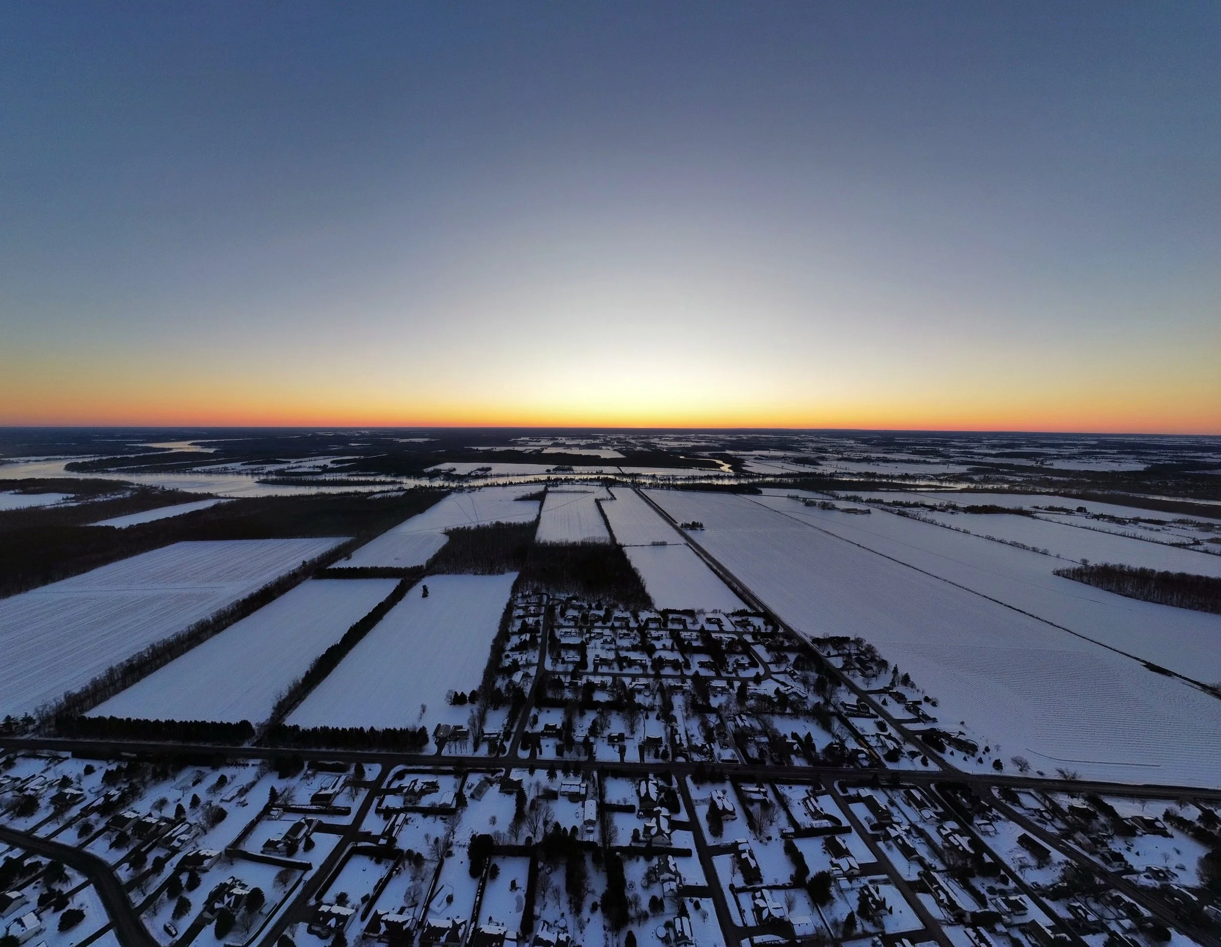 A winter landscape showing a flat area with snow-covered fields, roads, and houses, under a clear sky at sunset with a gradient from yellow to blue.