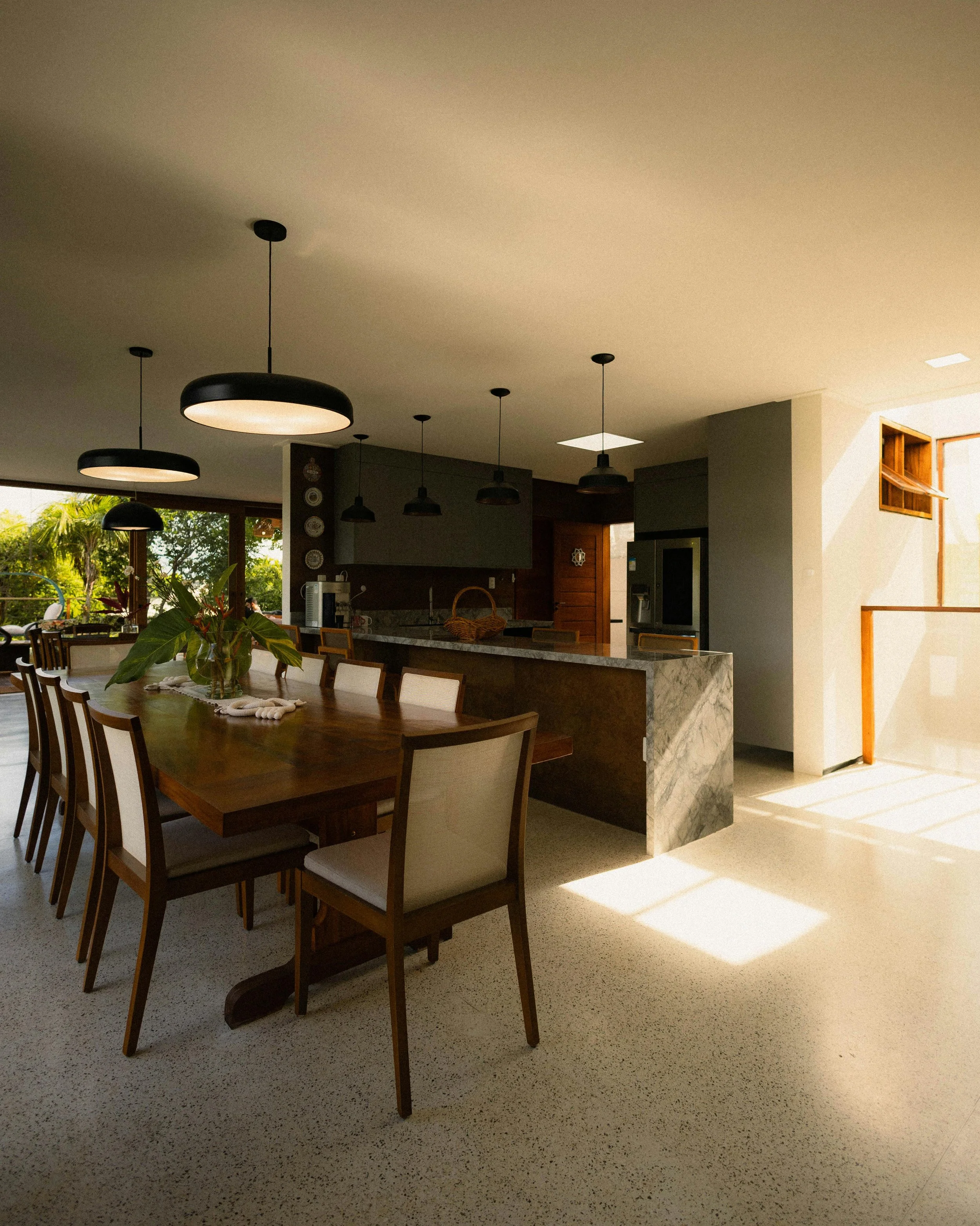 Interior of a modern house with a dining area in the foreground, featuring a wooden table with chairs and a plant centerpiece. In the background, there's a kitchen with a marble island, hanging pendant lights, and a sunny window showing greenery outside.