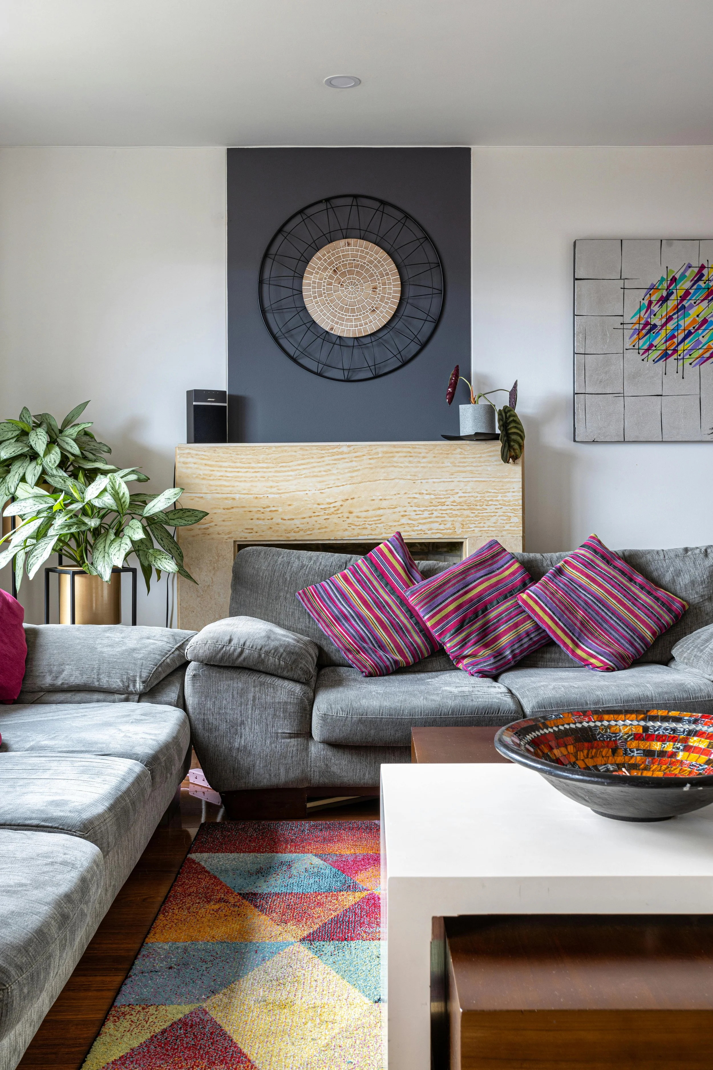 Living room with a grey sectional sofa adorned with colorful striped pillows, a wooden cabinet with a potted plant on top, a multicolored geometric rug, a white coffee table with a decorative bowl, a large abstract art piece, and a wall clock mounted on a dark accent wall.