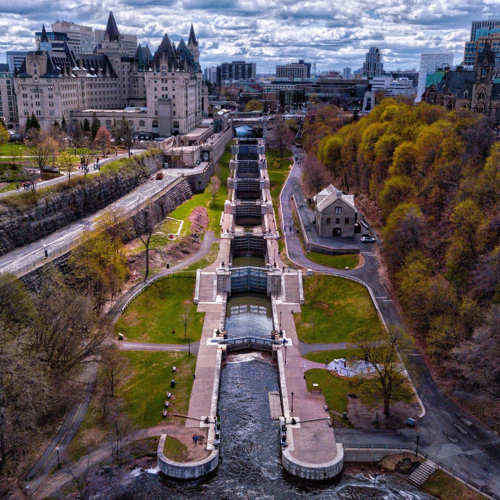 A cityscape featuring a historic castle-like building and a park with a cascading water feature in the foreground, with modern buildings and a cloudy sky in the background.