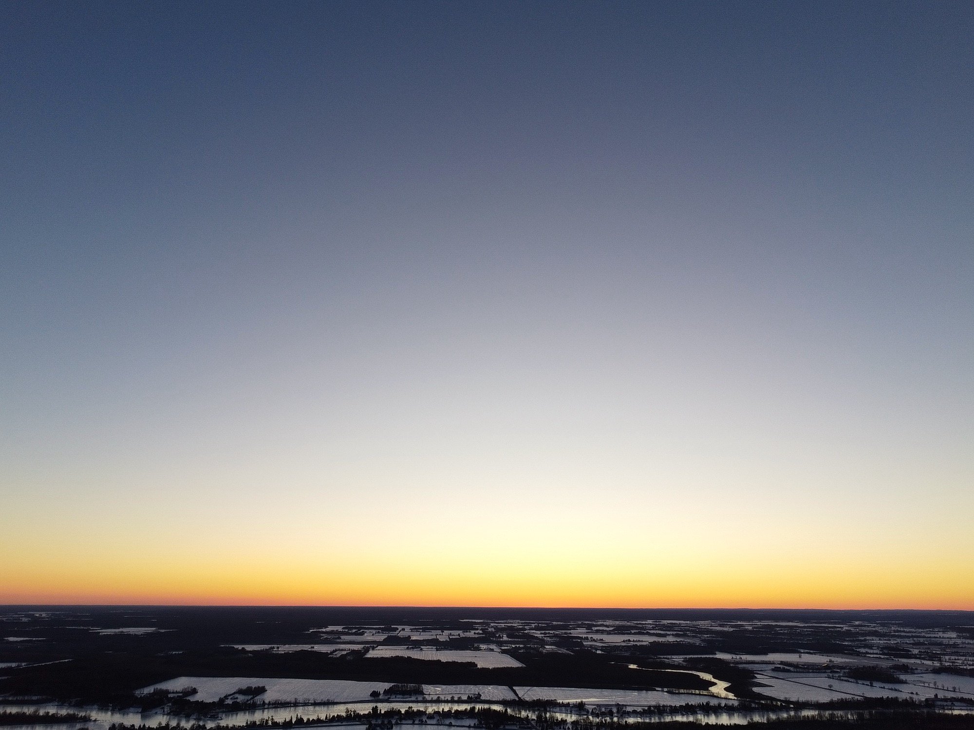 A wide shot of a winter landscape at sunset or sunrise, with snow-covered fields, patches of water, and sparse trees, under a sky shifting from yellow near the horizon to blue higher up.