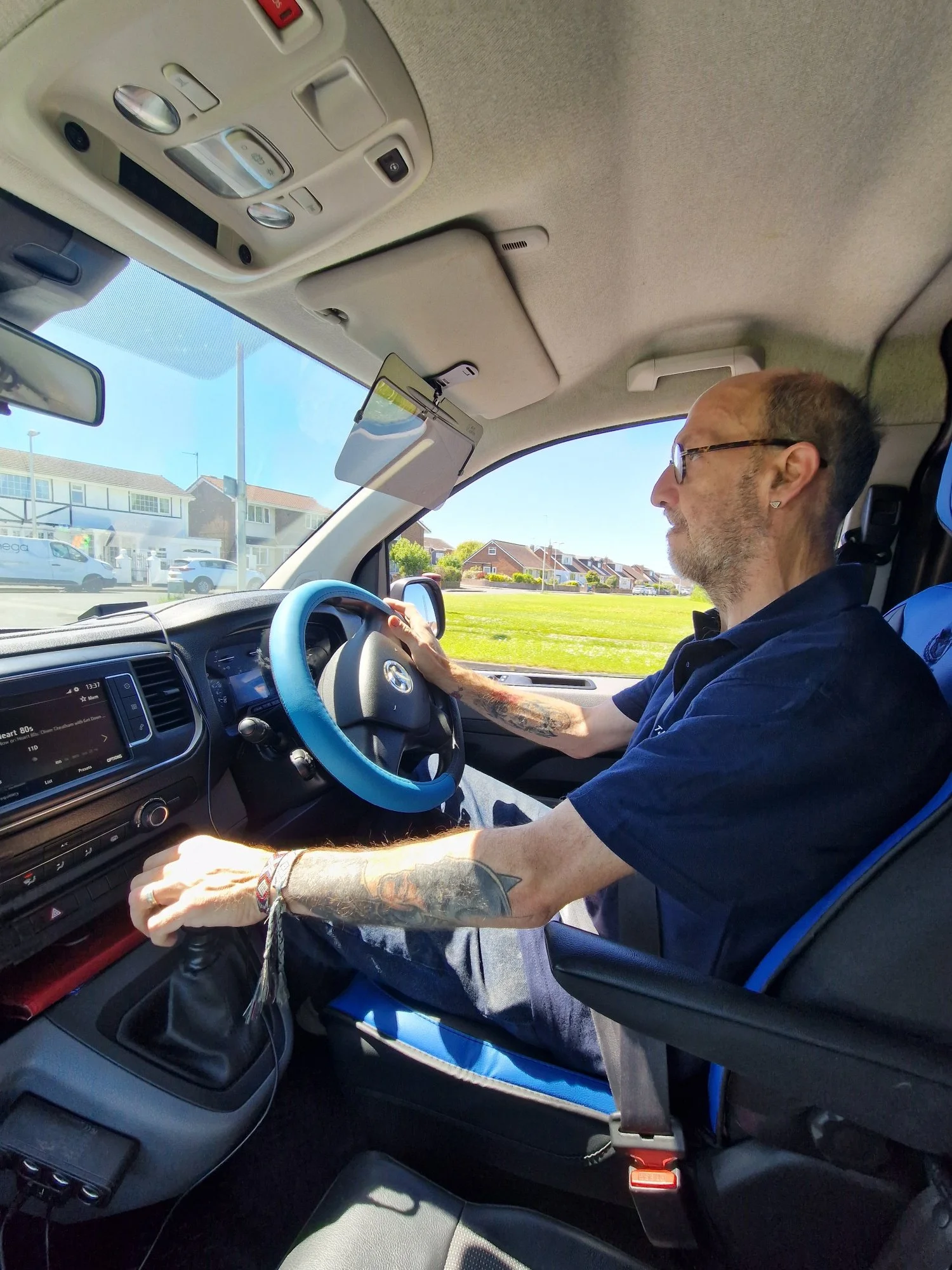 A man with tattoos on his arm sitting in the driver's seat of a vehicle, wearing glasses, with his hand on the steering wheel, in a bright, sunny environment with houses visible outside.