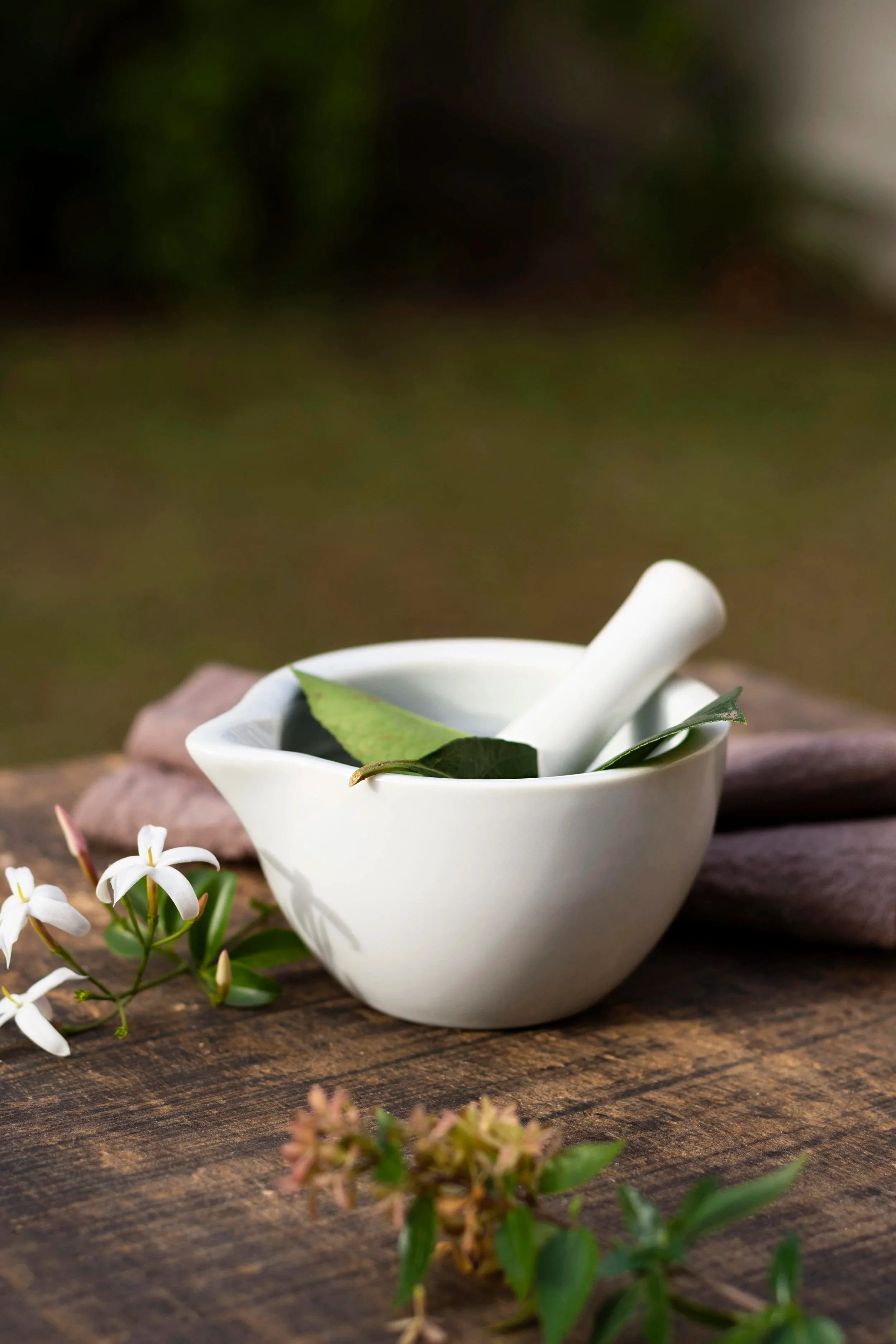 A white ceramic mortar and pestle with green leaves inside, placed on a rustic wooden surface, with small white flowers and pinkish-red buds nearby, blurred background of greenery.