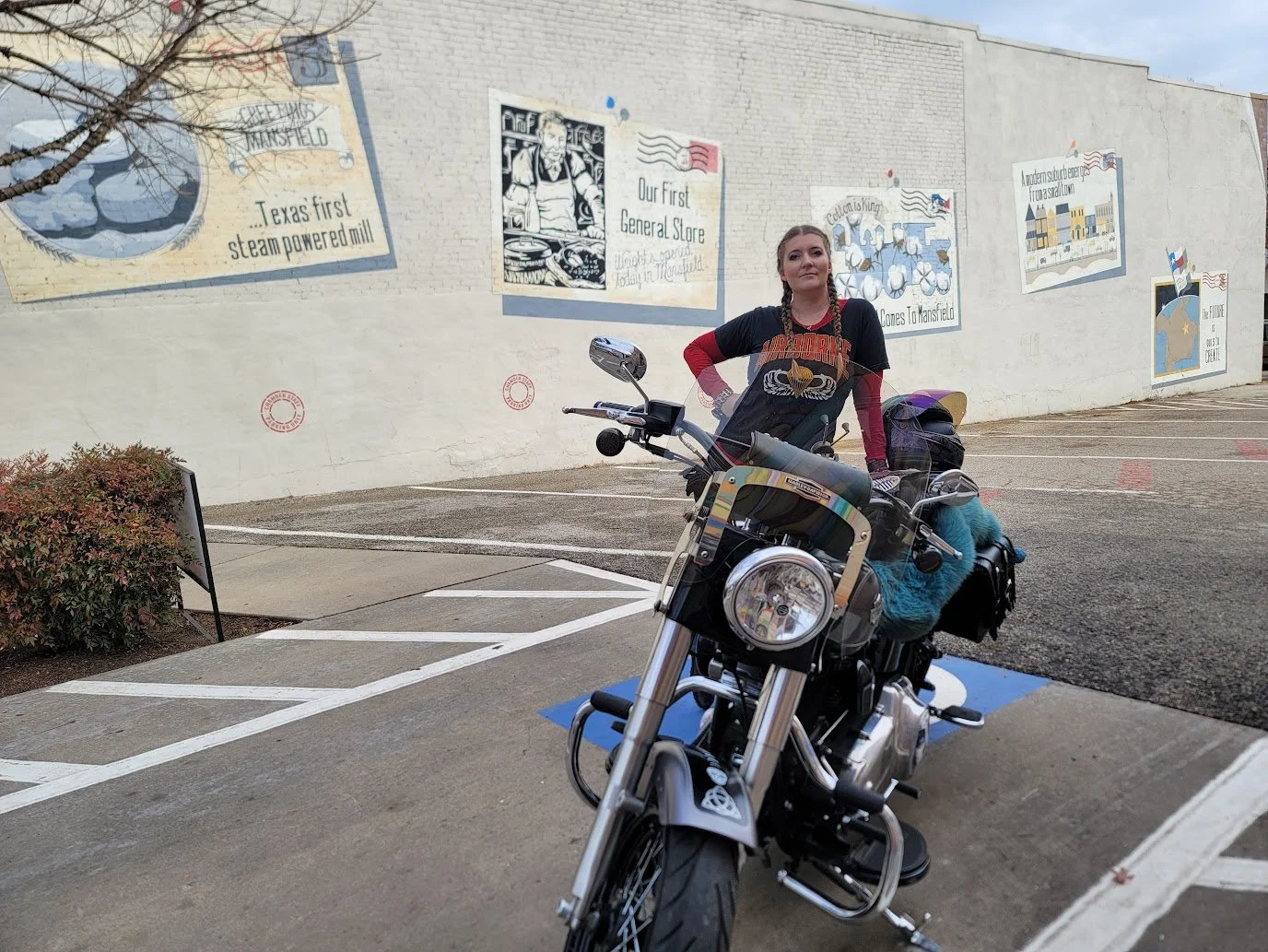 A woman standing next to a black motorcycle in a parking lot with a mural on the wall behind her.