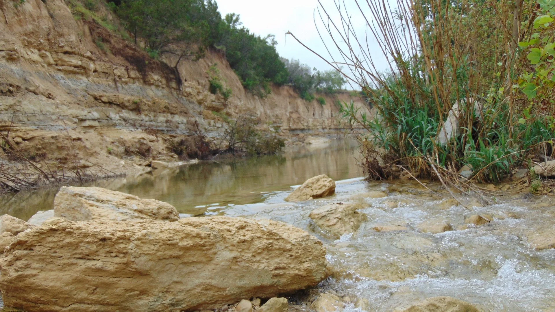 A small river or creek flowing through a rocky landscape with steep, eroded cliffs on one side and tall green grass and trees on the other.