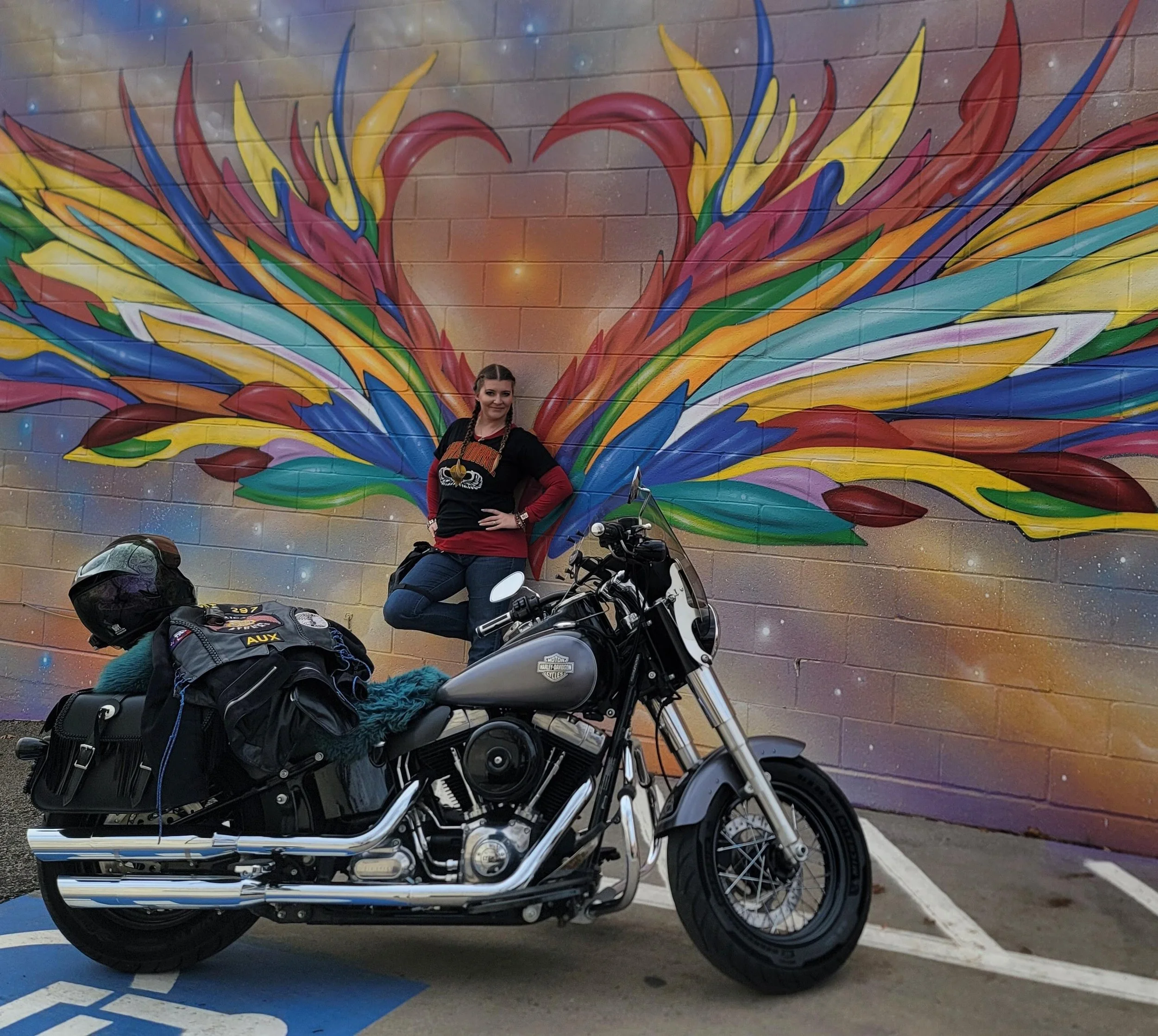 A woman standing next to a parked black Harley-Davidson motorcycle in front of a colorful mural on a brick wall, with parts of helmets and motorcycle gear in the foreground.